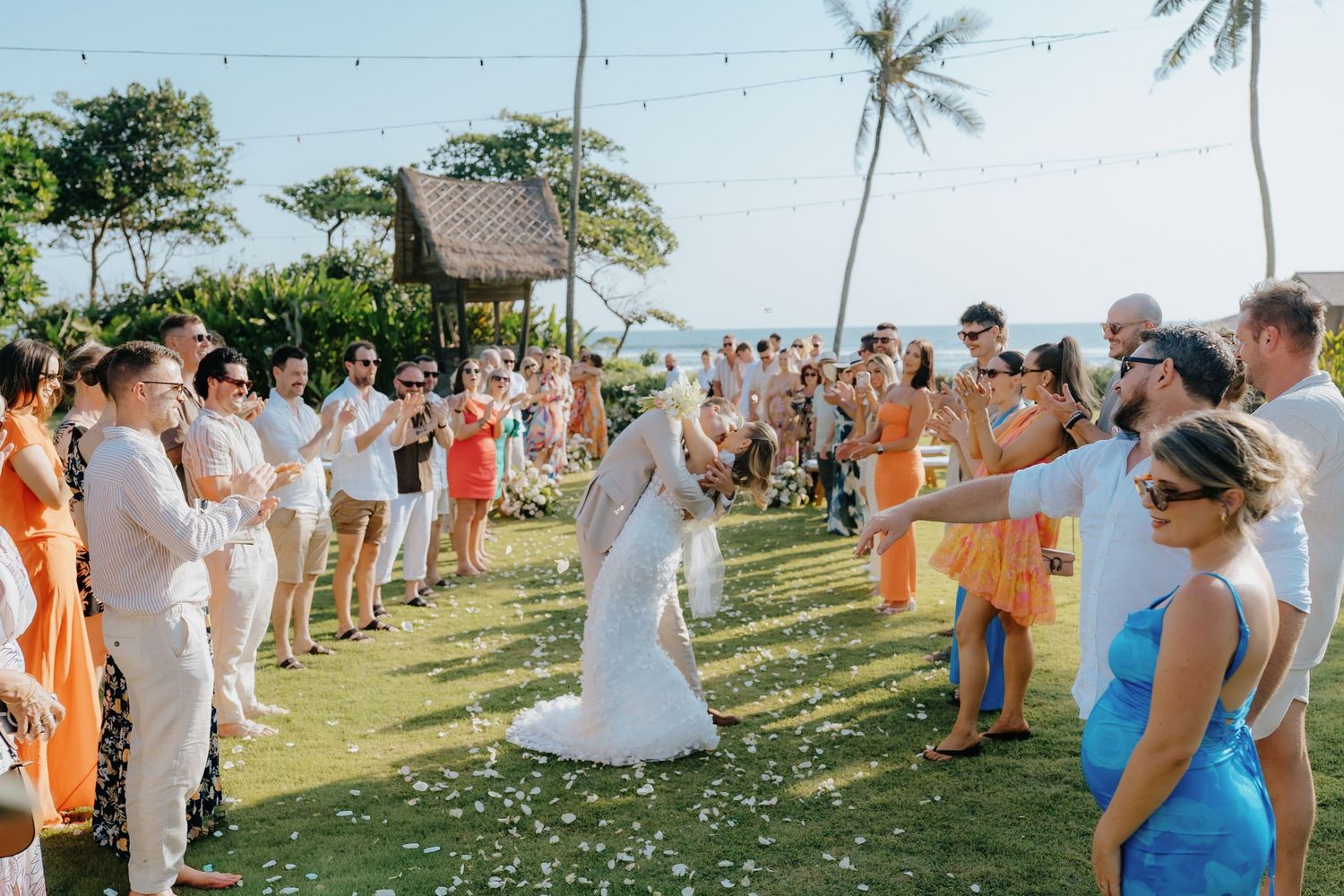 Bride and groom walk through flower petal aisle at outdoor tropical beach wedding ceremony with guests cheering.