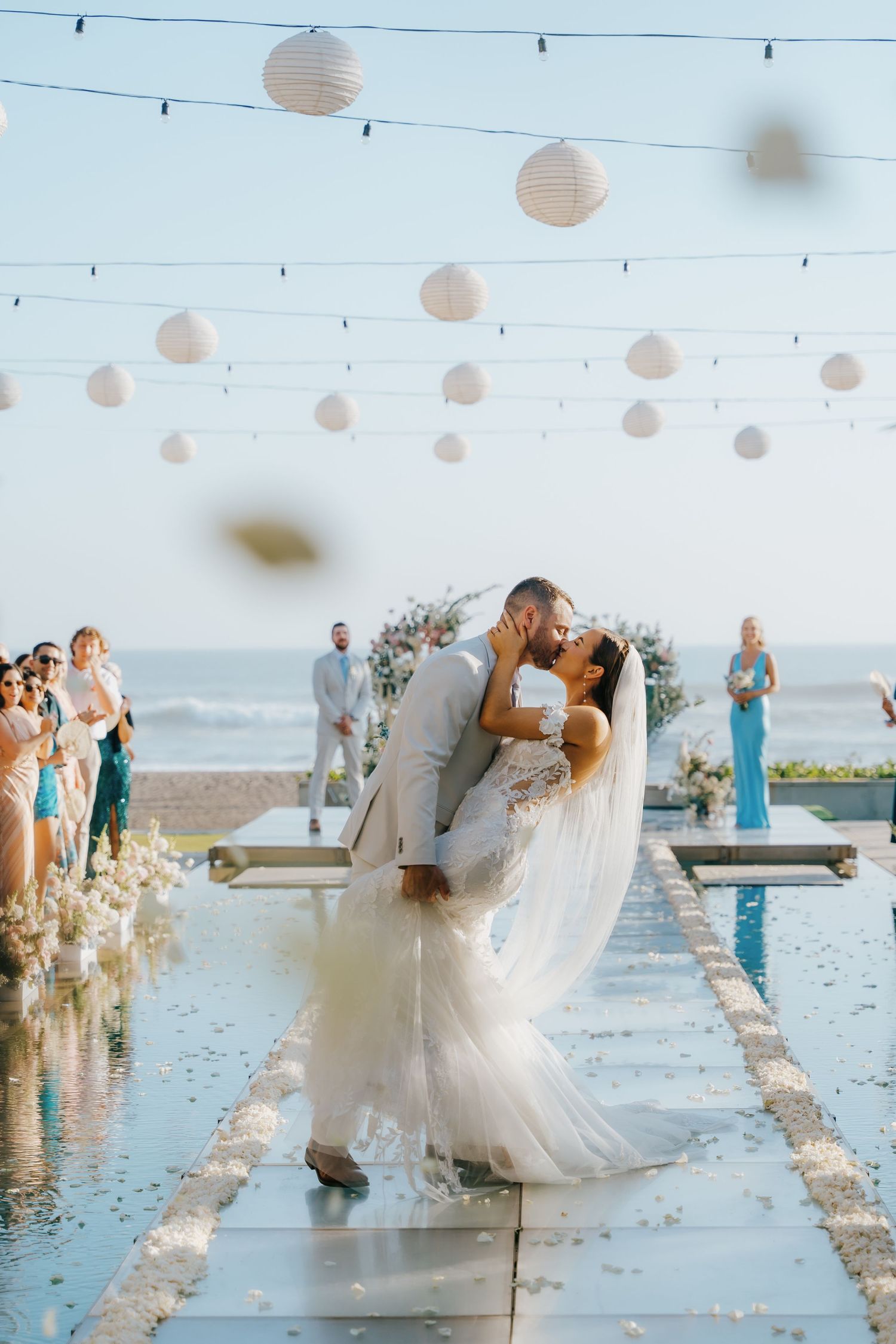 Bride and groom share a romantic dip kiss on a mirrored aisle, surrounded by white lanterns and ocean views.