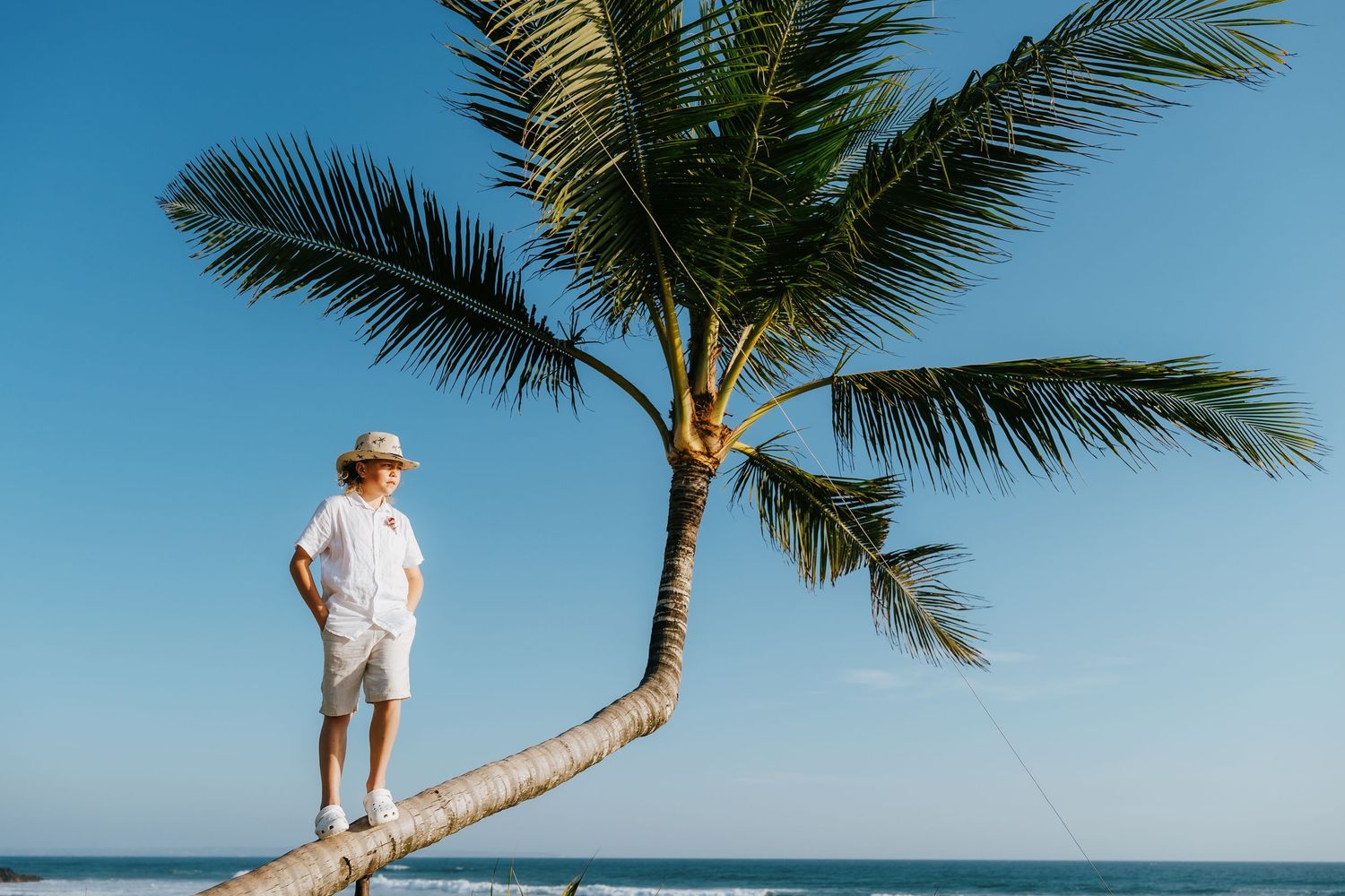 Child in white outfit and hat standing on a leaning palm tree trunk at a tropical beach with clear blue sky.