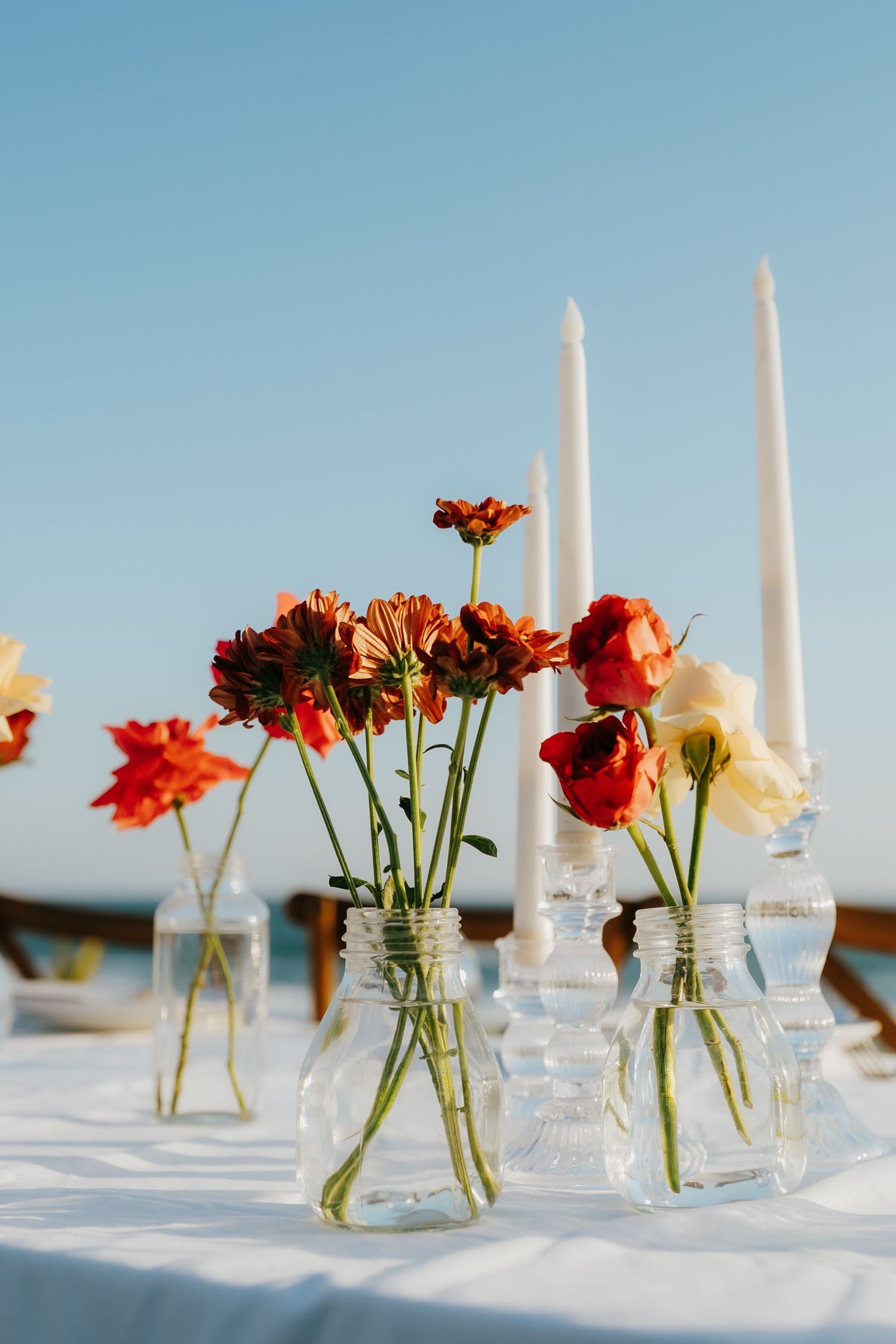 Red and yellow flowers in glass mason jar vases with white taper candles on a white tablecloth outdoors.