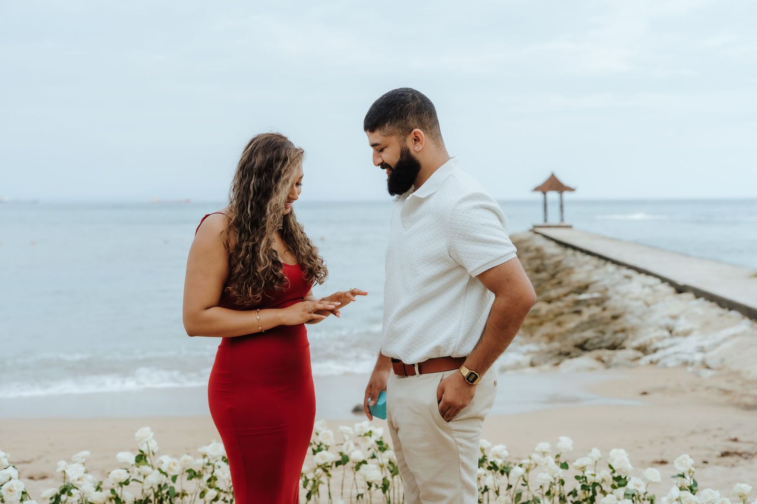 A couple stands on a tropical beach walkway, with the woman in a red dress and man in white against an ocean backdrop.