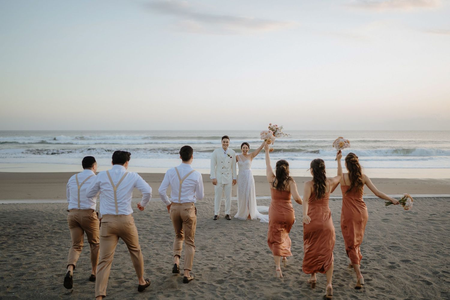 Wedding party running toward bride and groom on a sandy beach at sunset with ocean waves in the background.