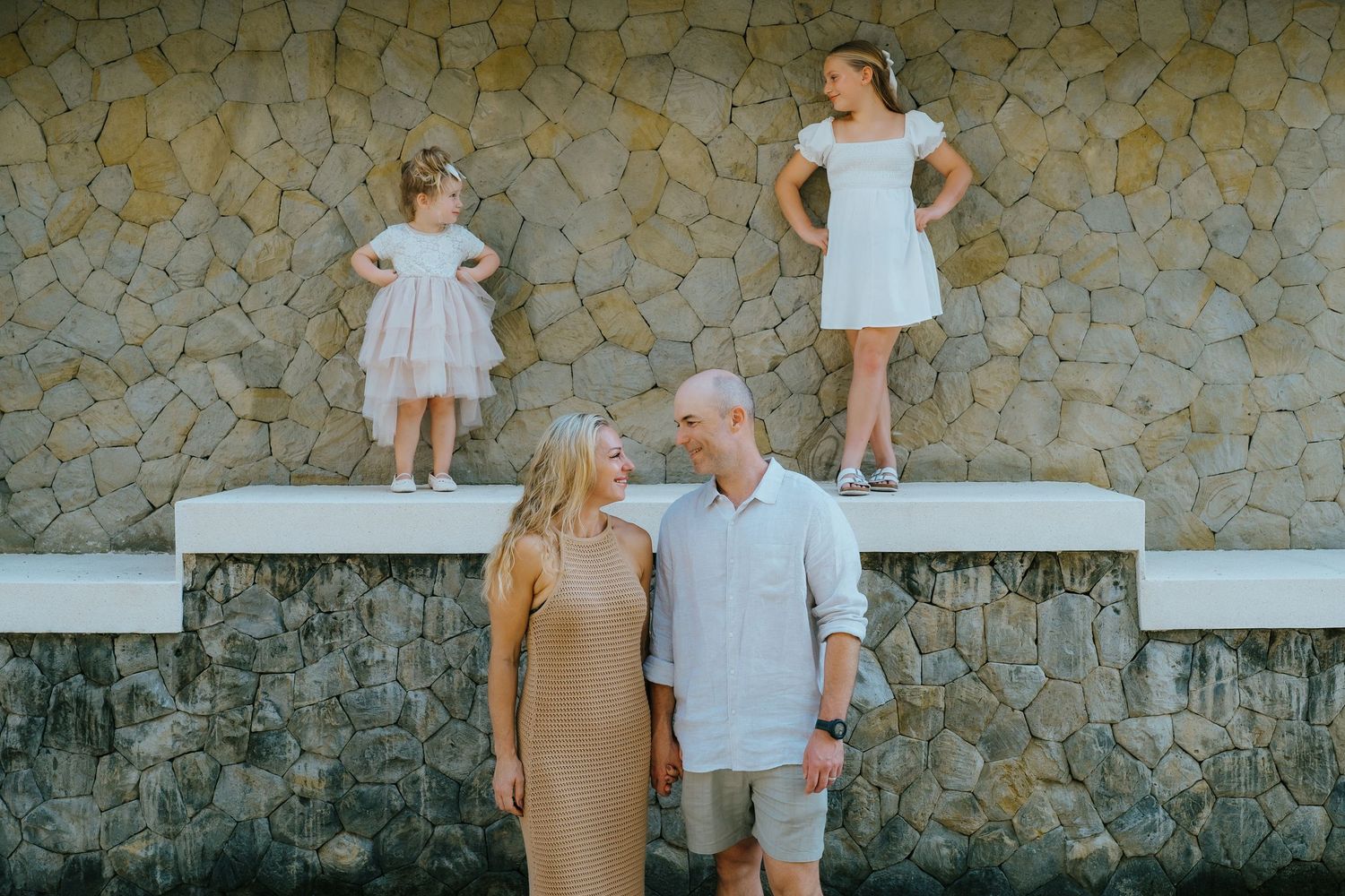 Happy family with two young girls standing on stone wall, smiling parents in foreground at outdoor photo session.