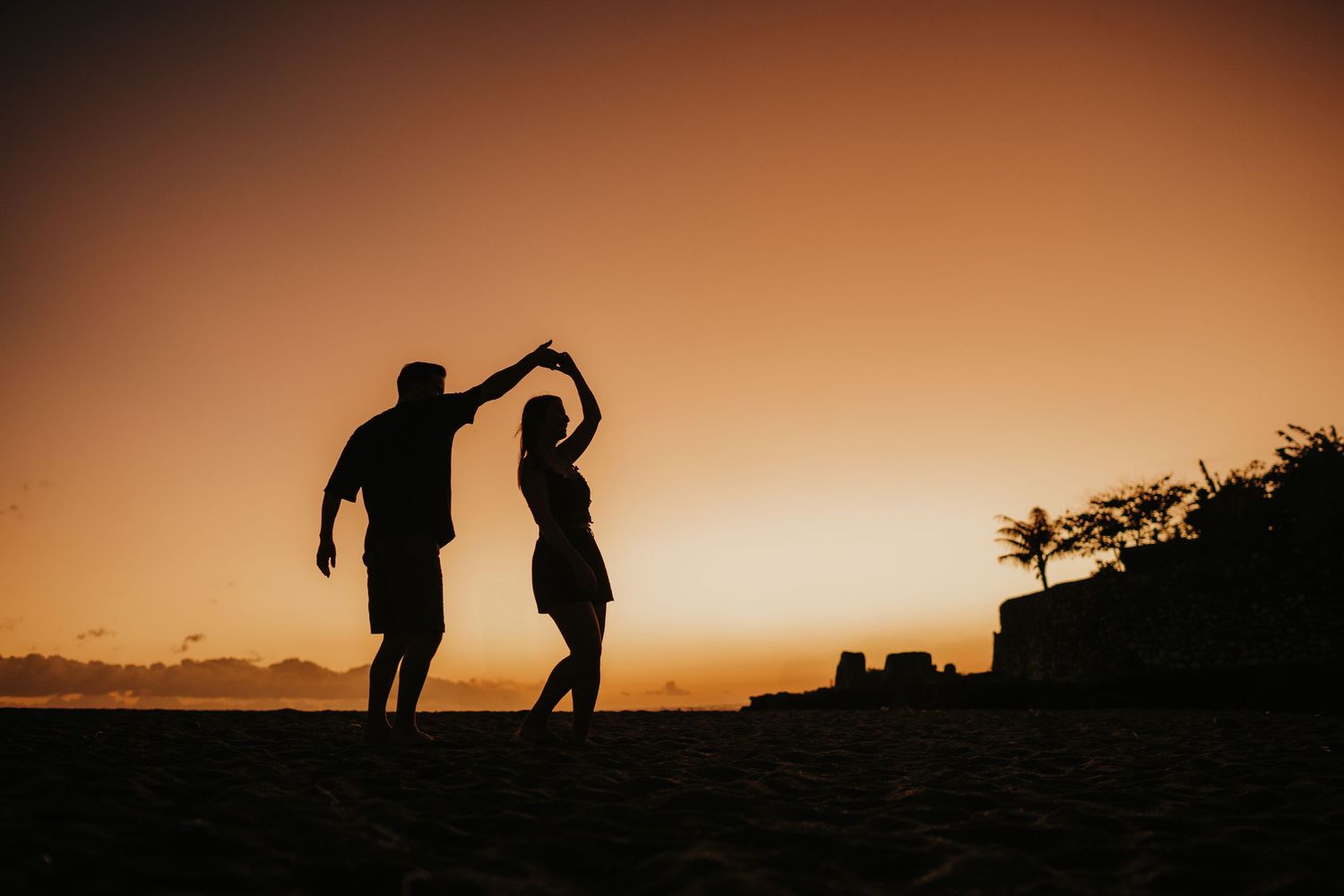 Silhouettes of couple dancing and playing against dramatic orange sunset sky.