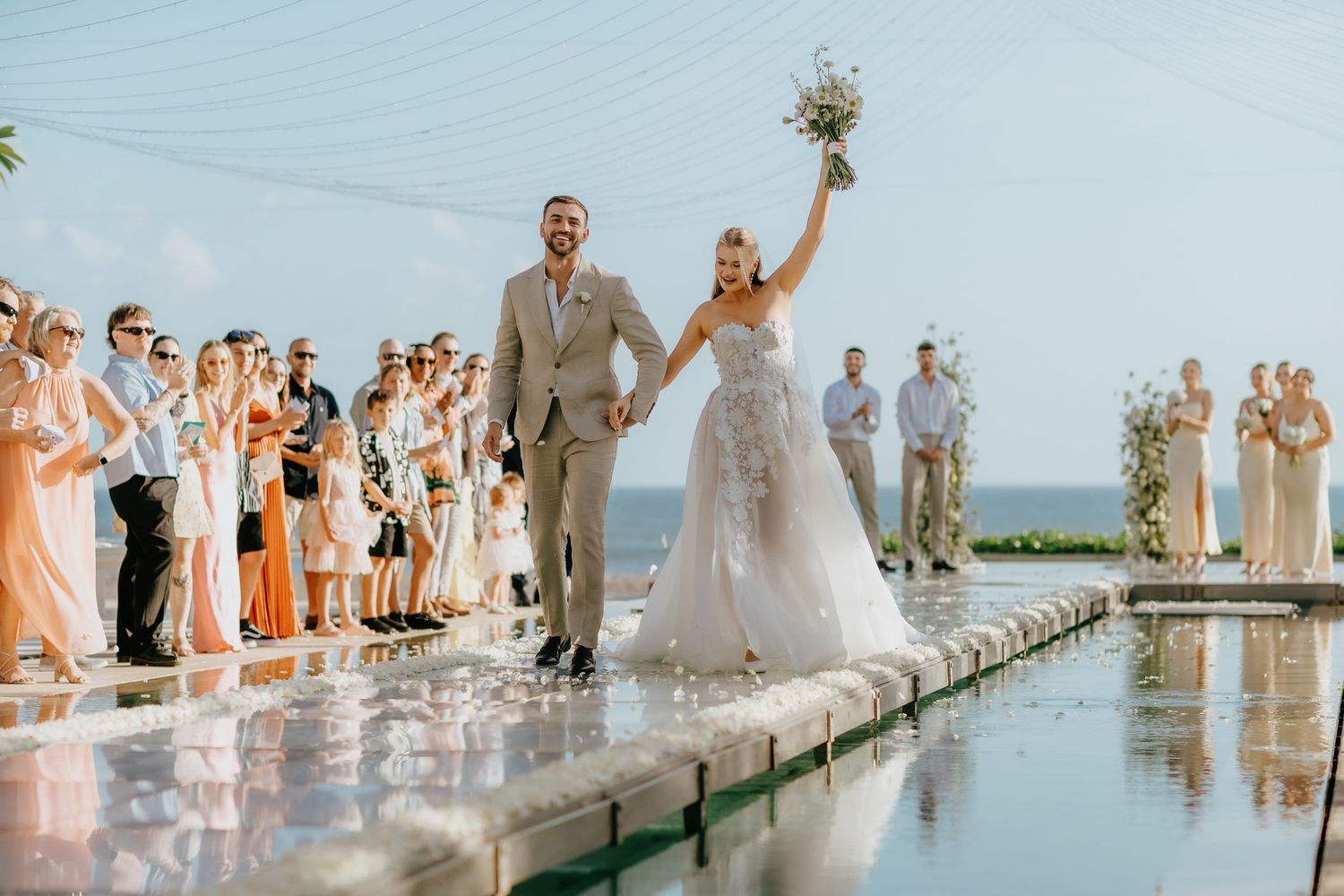 Bride and groom walk down aisle over reflective pool at outdoor wedding ceremony with ocean view.