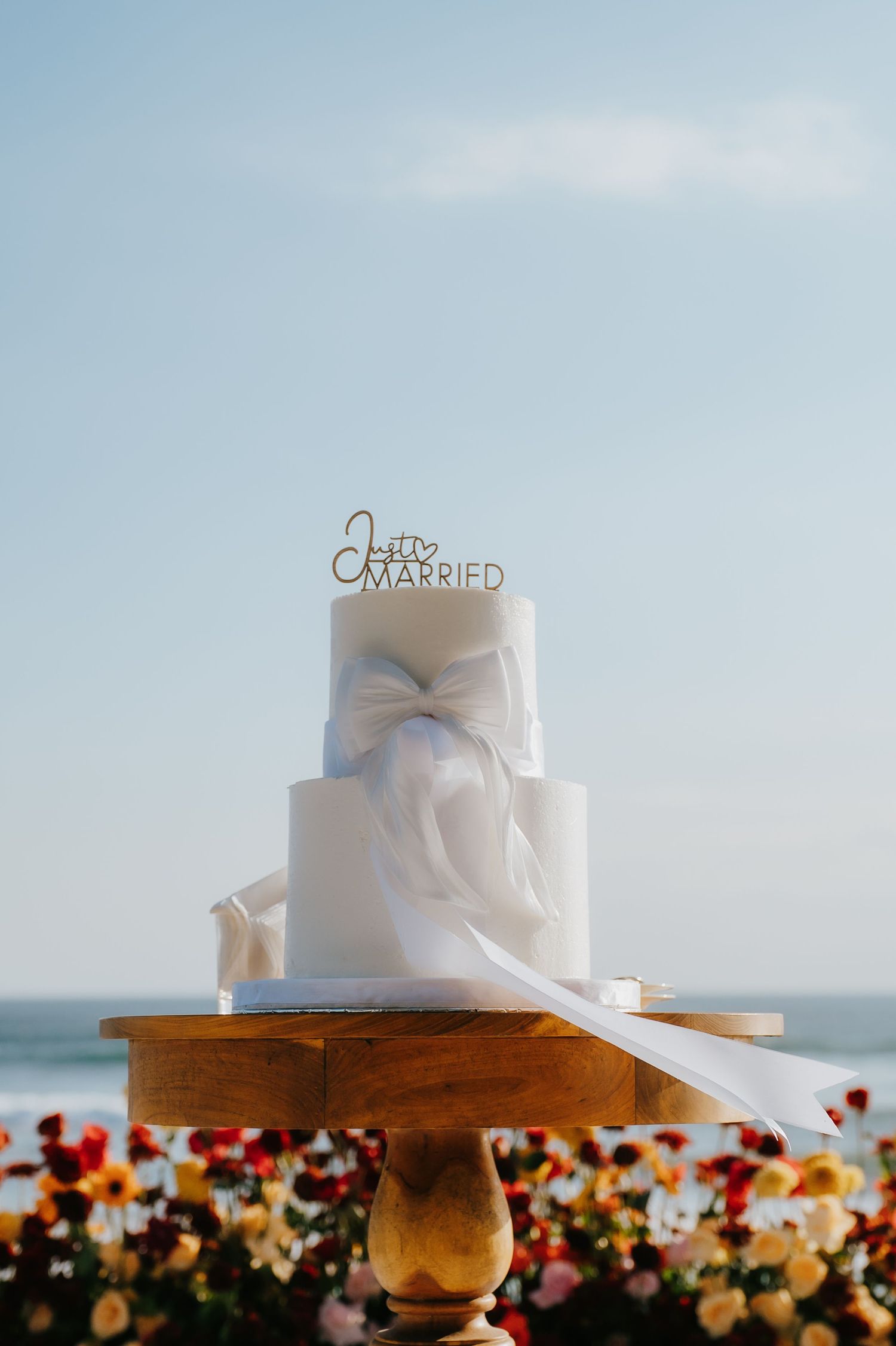 Three-tier white wedding cake with bow and Just Married topper on wooden stand, ocean backdrop with colorful flowers.