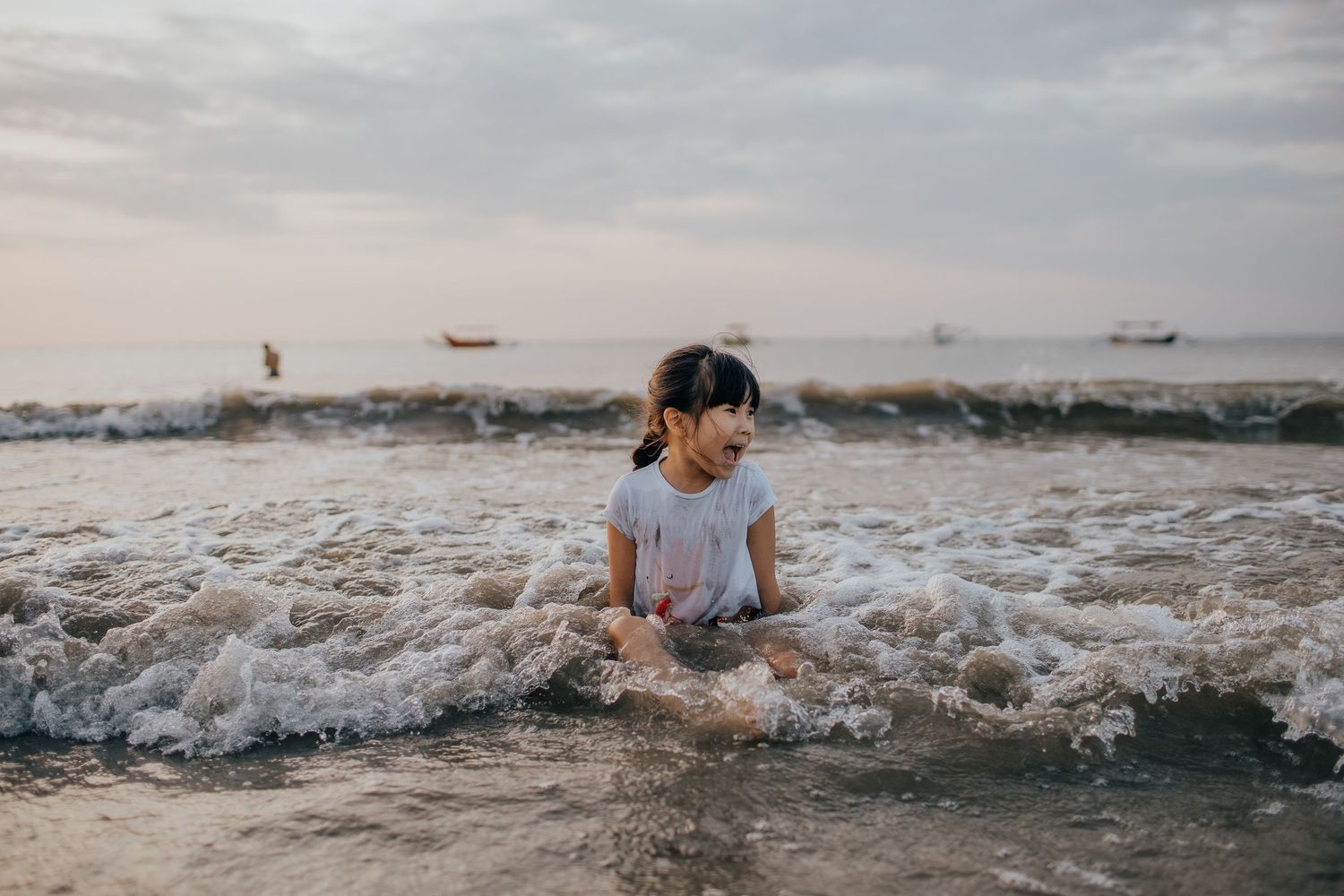 Child sitting in ocean waves at sunset with boats visible on horizon.