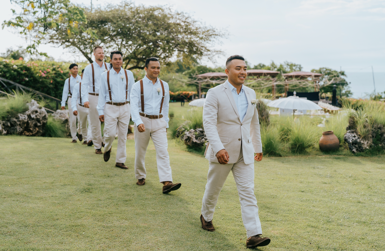 Groom leads groomsmen in white suits and suspenders across green lawn at outdoor wedding venue.