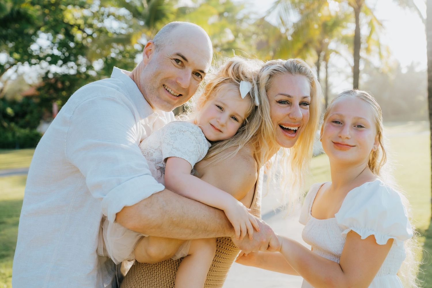 Happy family of four posing outdoors at sunset with palm trees, featuring a bald man, blonde woman, and two young girls.