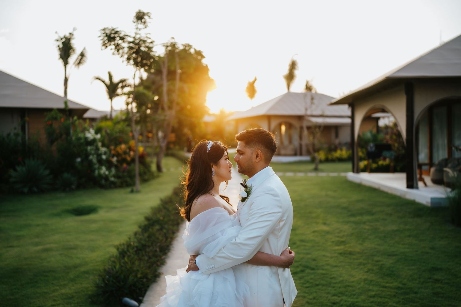 Bride and groom share a romantic moment at sunset in a lush garden with tropical villas in the background.