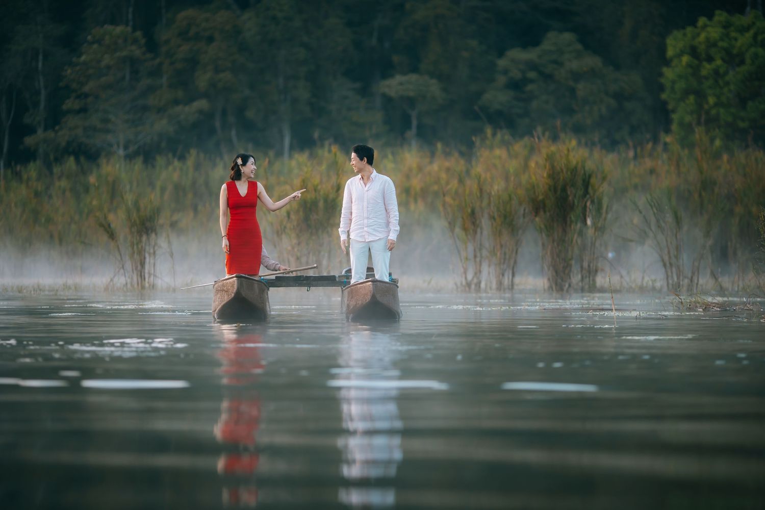 Couple standing on wooden boats in misty lake surrounded by lush greenery, woman in red dress, man in white outfit.