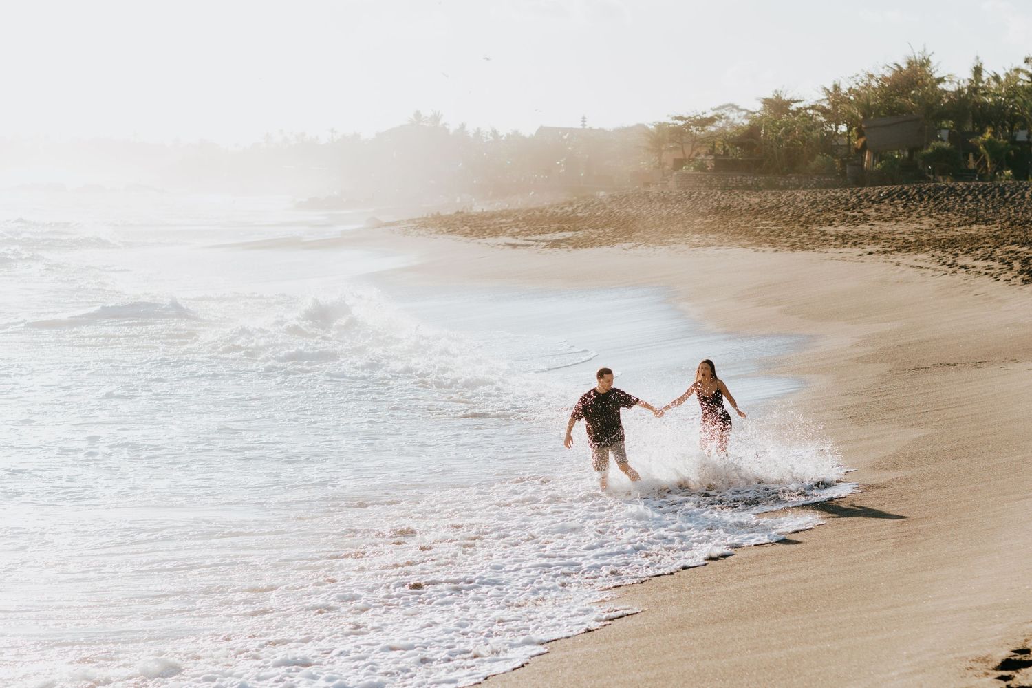 Two people playfully run through shallow ocean waves on a tropical beach at sunset.