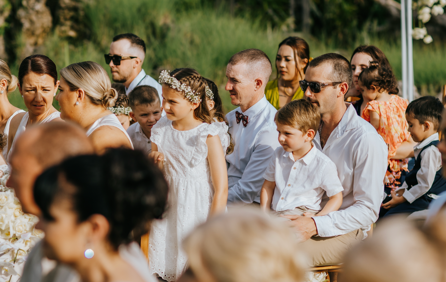 Children and adults in formal attire attend an outdoor wedding ceremony, with flower girls in white dresses standing at the front.
