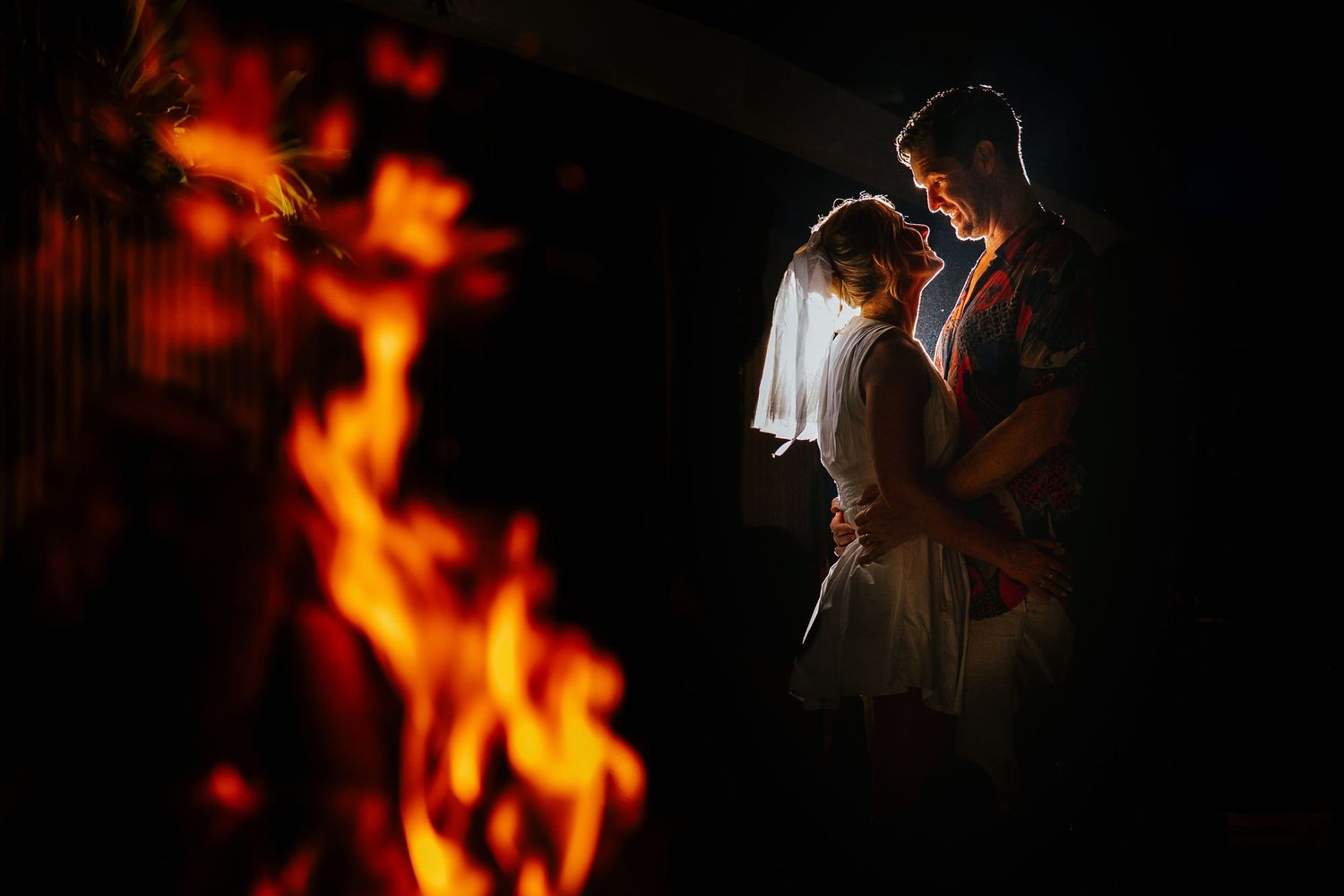 Romantic wedding couple embracing by firelight in a dark, dramatic setting, bride in white veil glowing warmly.