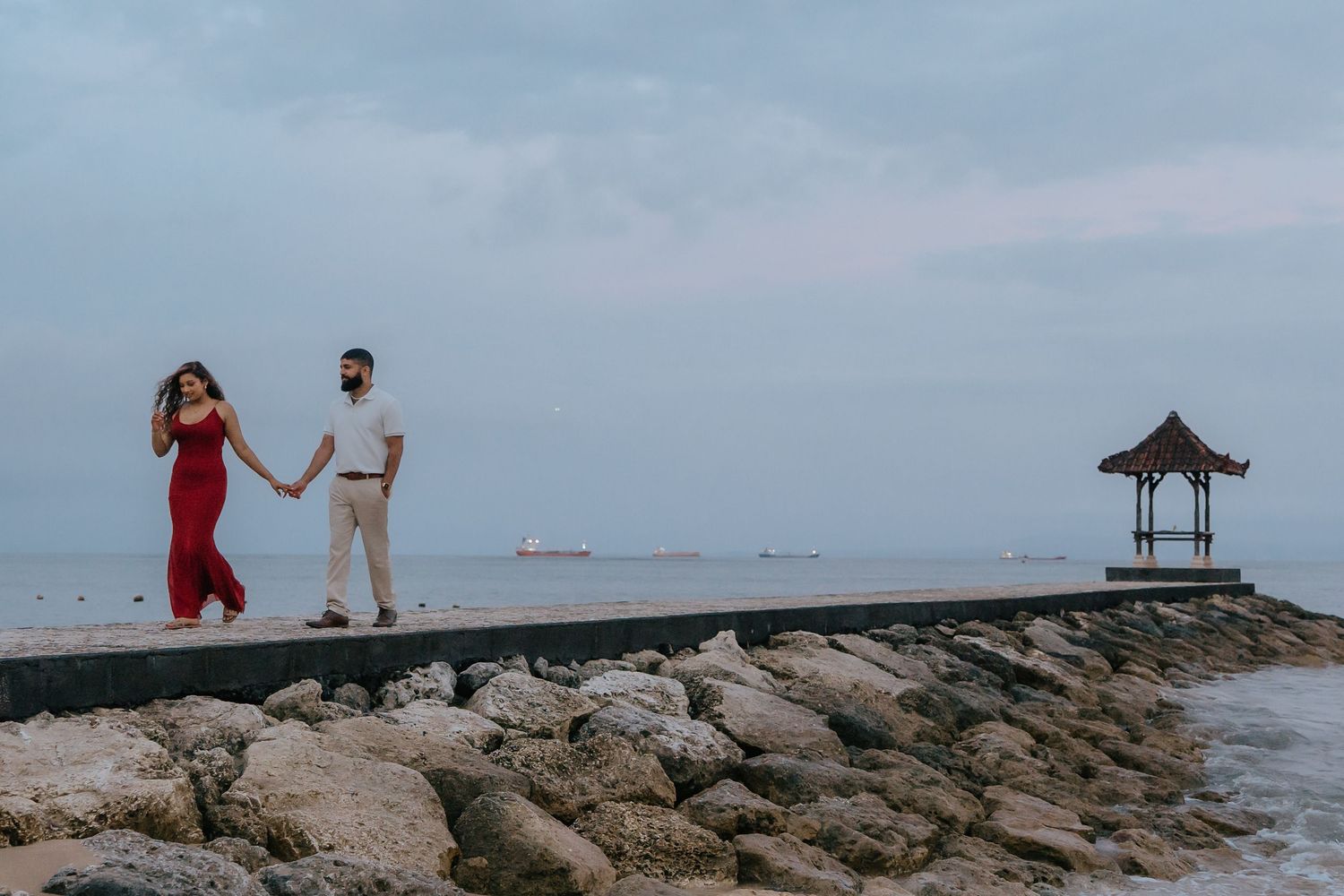 People walk along a rocky beach jetty overlooking the ocean on a cloudy day.