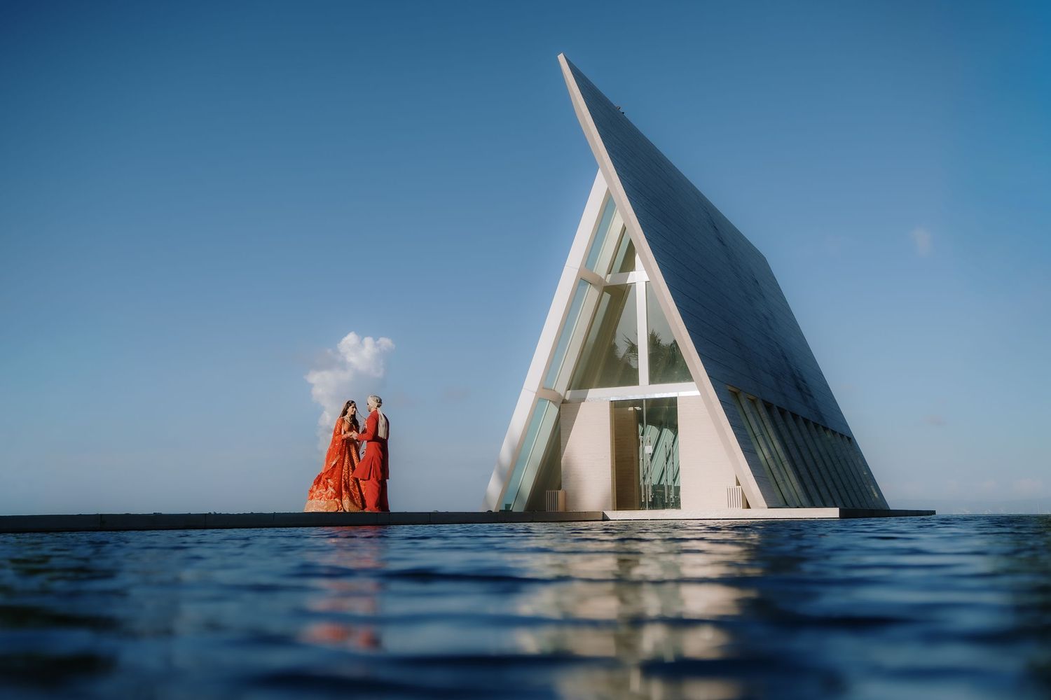 Two women in orange dresses stand near a modern triangular white chapel partially submerged in calm blue water.
