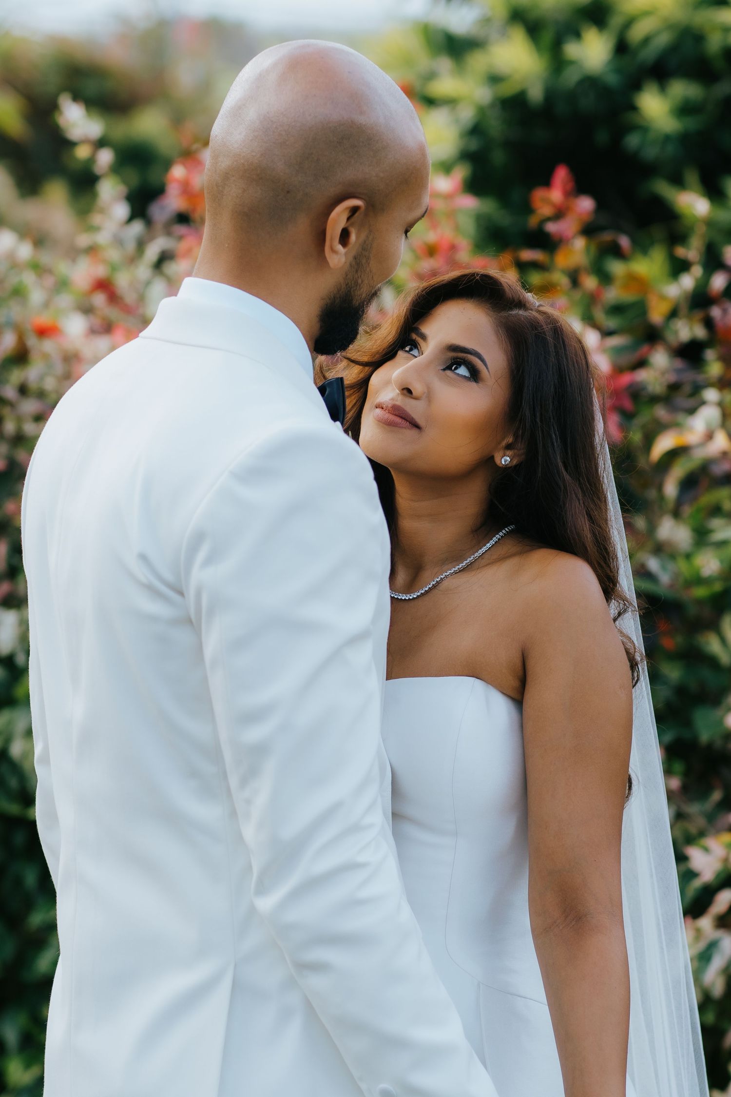 Bride in strapless white gown gazes up at groom in white suit, surrounded by lush floral greenery.