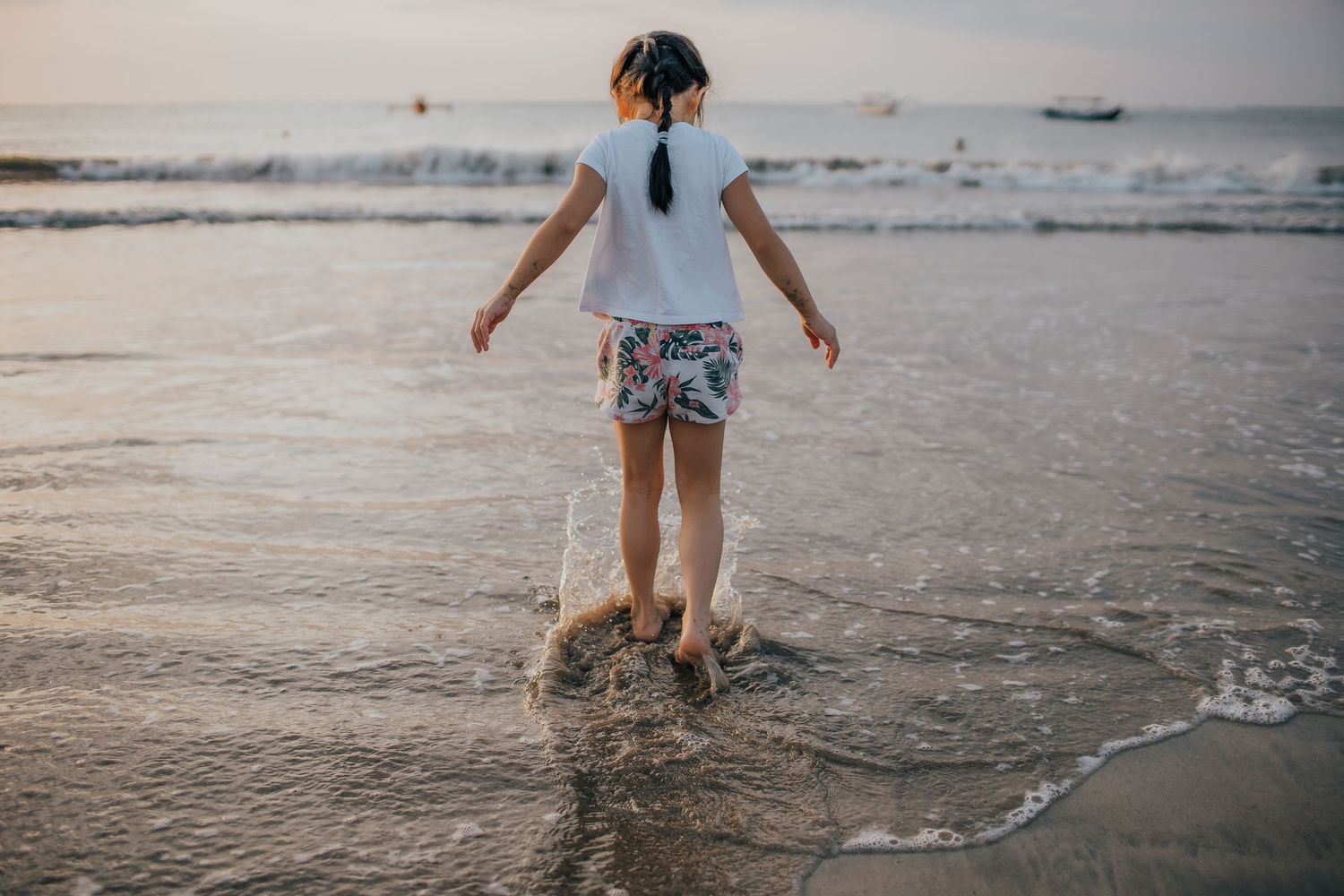 Child splashing in shallow ocean waves while wearing white shirt and patterned shorts.