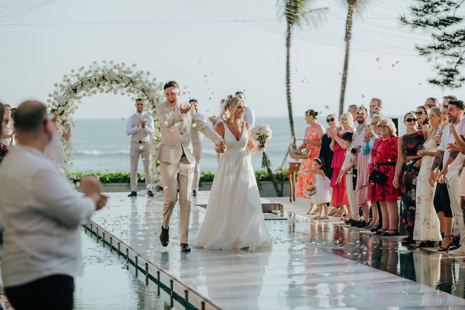 Newlywed couple kissing at outdoor beach wedding ceremony with floral arch and guests celebrating.