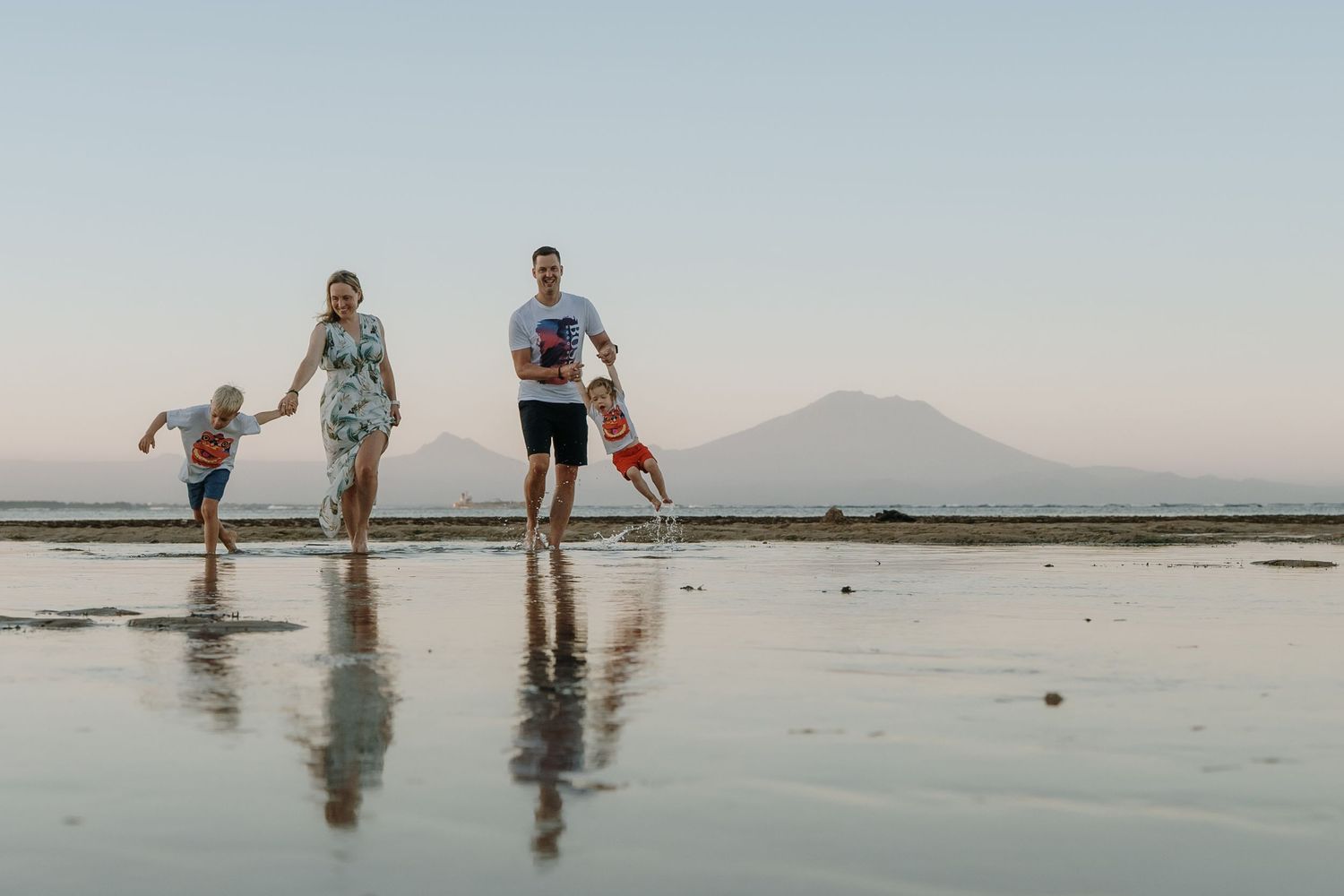 Family running joyfully on a reflective beach at sunset with a mountain backdrop.