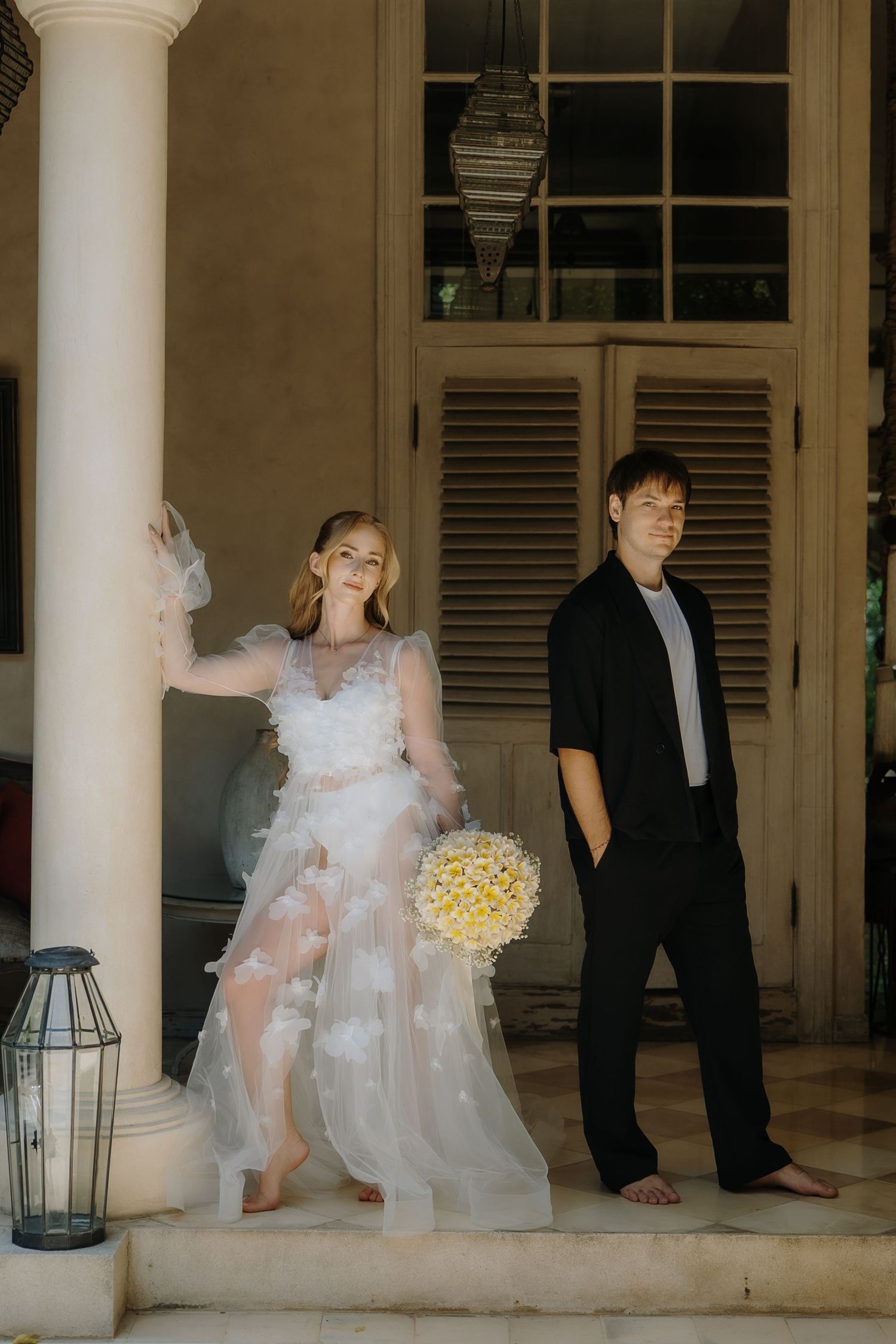 Bride in sheer white gown holding yellow bouquet poses with groom in black suit on elegant villa steps.