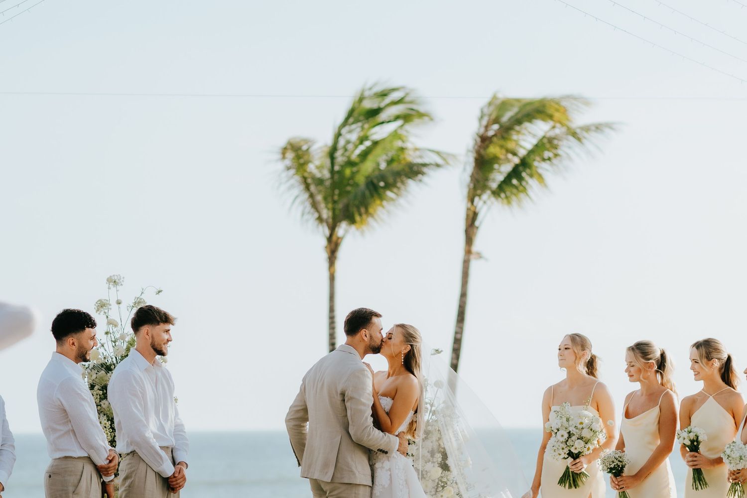 Bride and groom share a kiss at a tropical beach wedding ceremony with palm trees and bridesmaids nearby.
