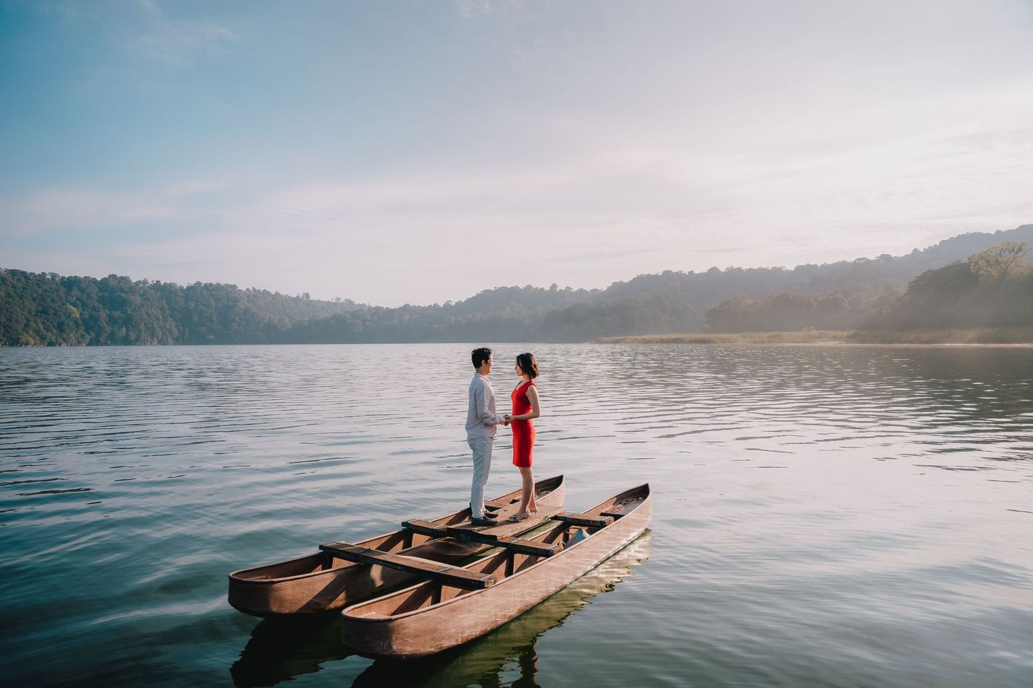 Couple standing on a wooden boat on a calm lake surrounded by lush green hills in soft morning light.