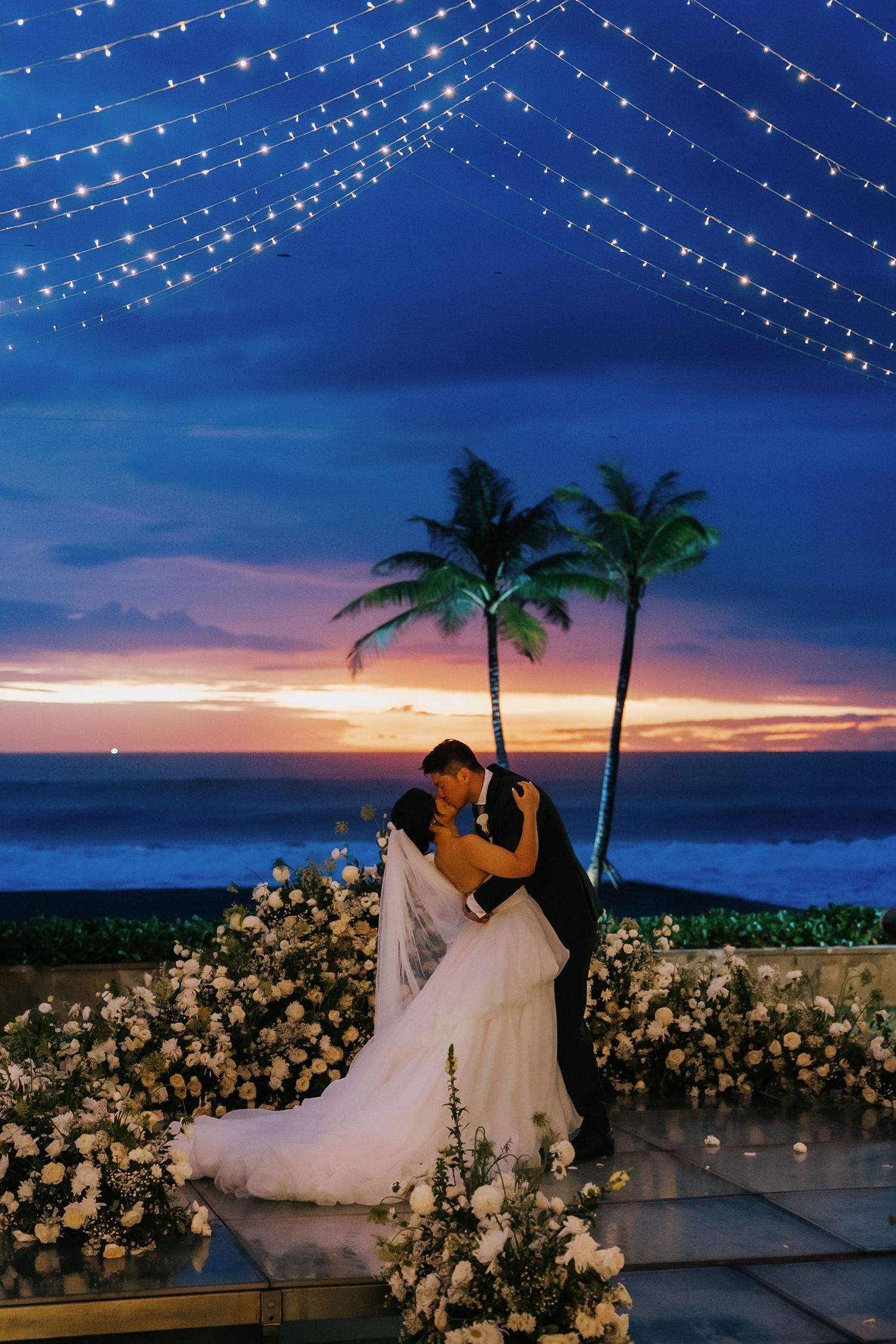 Couple shares a romantic kiss at sunset beach wedding with fairy lights and tropical palm trees backdrop.