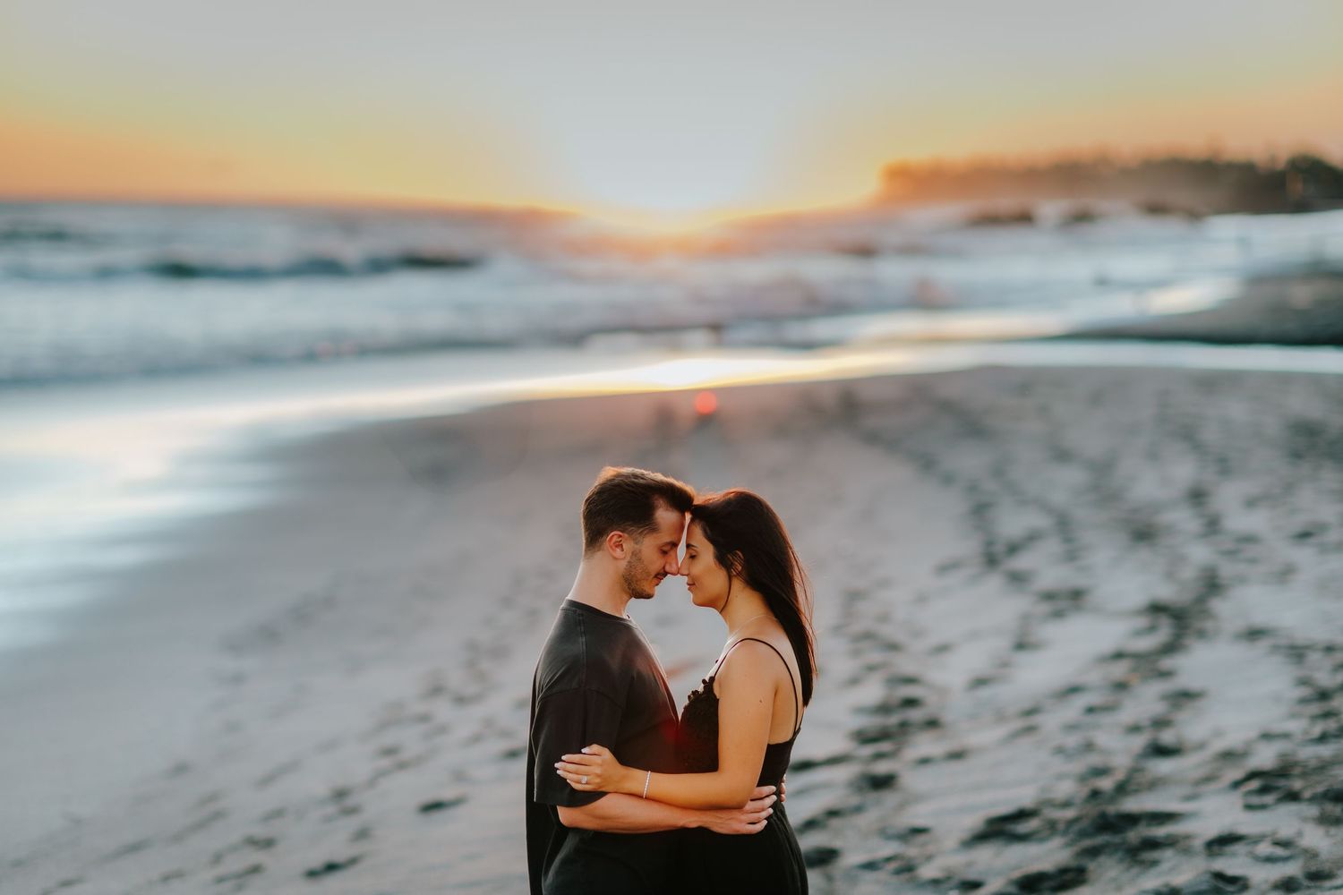 Romantic couple embraces on beach during golden hour sunset with waves crashing behind them.