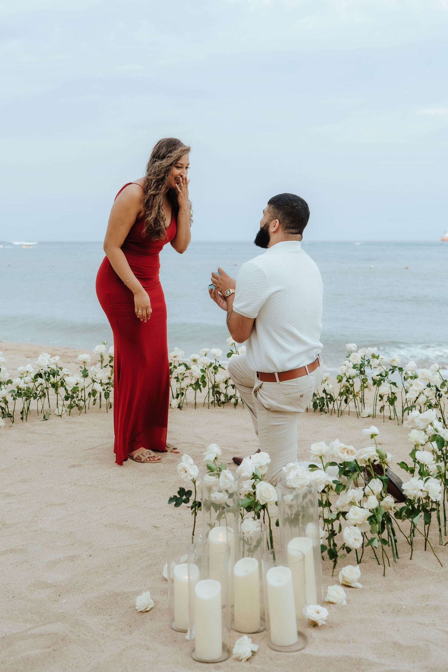 A romantic beach proposal scene with white candles and flowers arranged on the sand by the ocean.