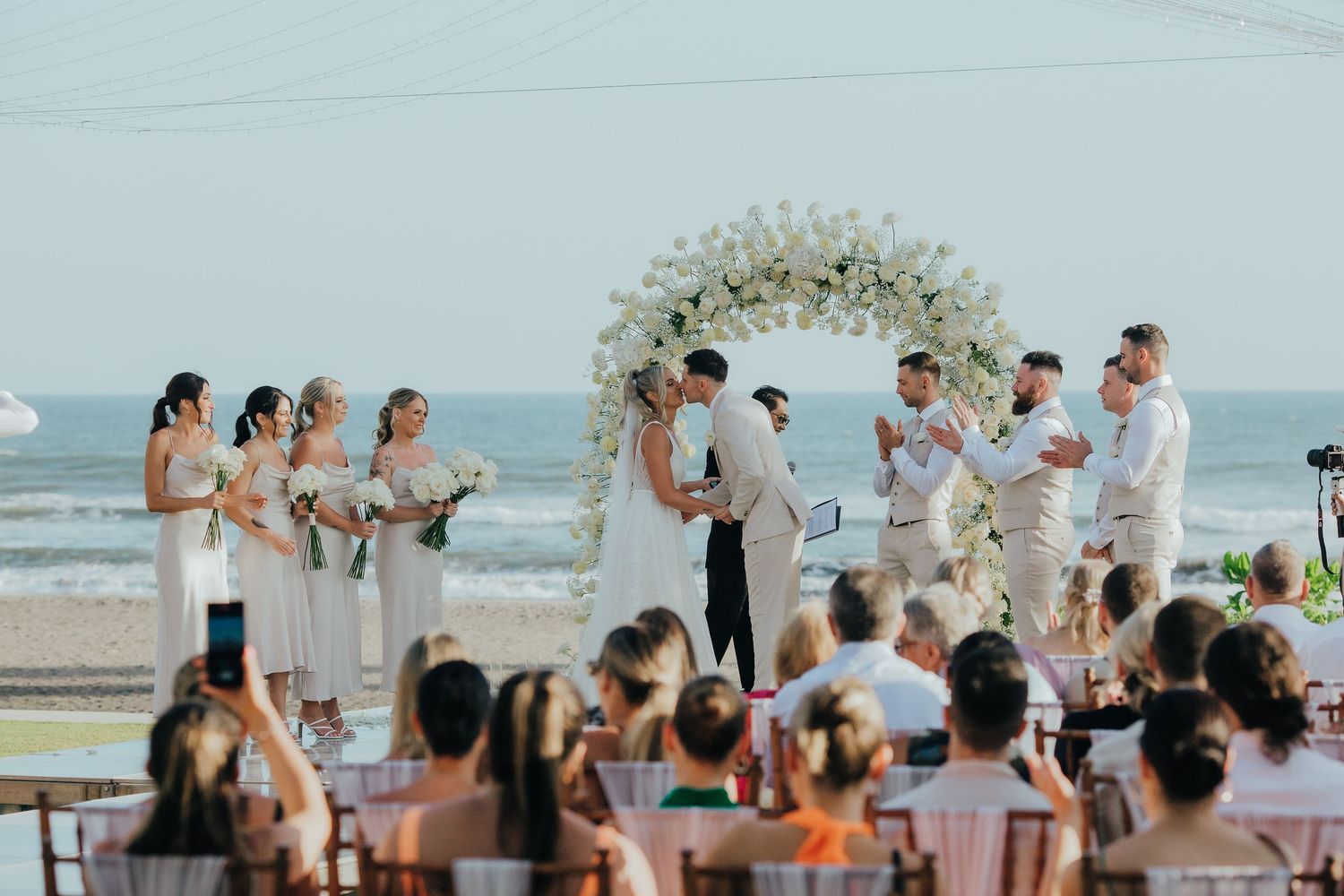 Beach wedding ceremony with couple kissing under floral arch, bridal party in white, ocean backdrop.