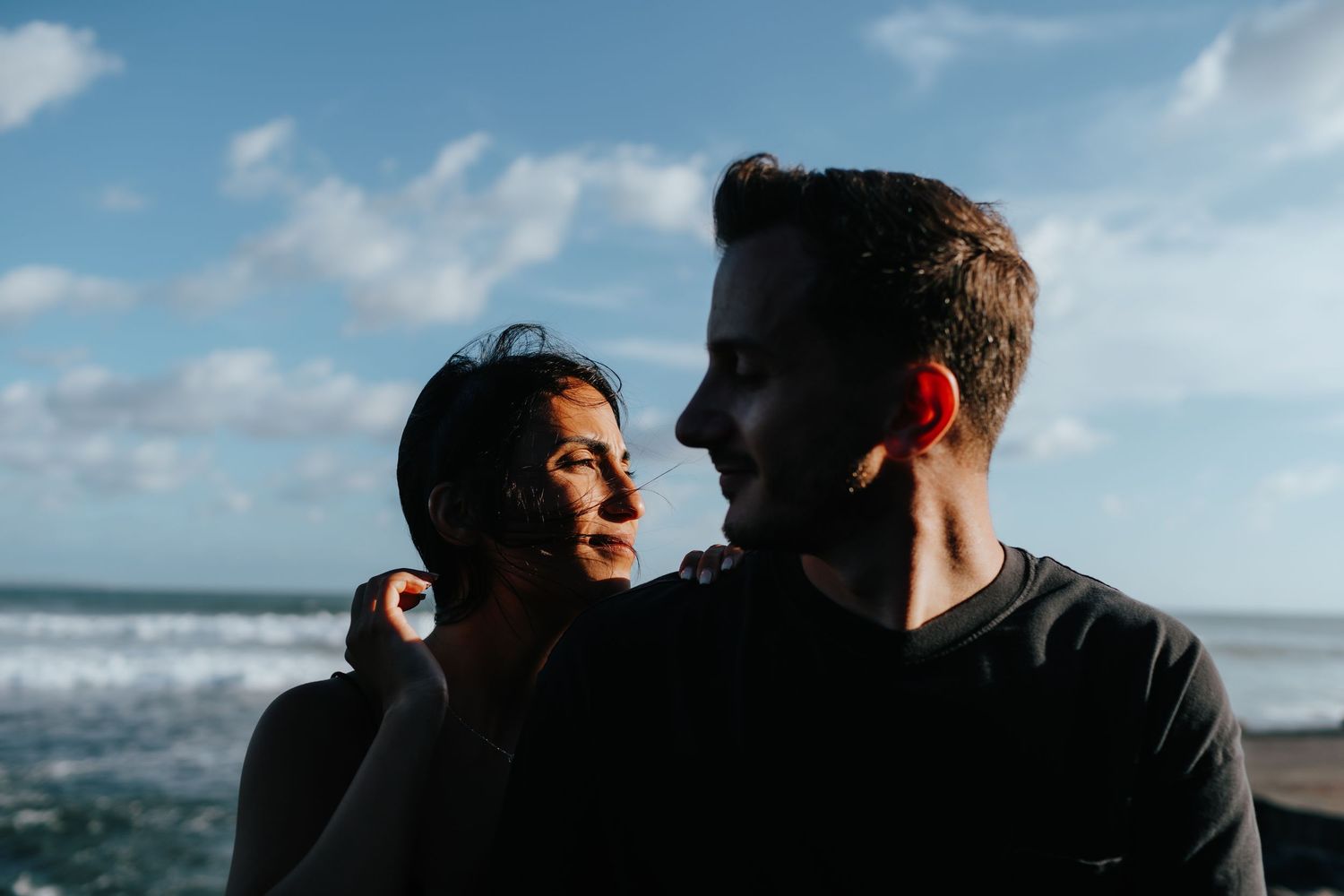 Couple's silhouettes against bright blue sky and clouds by the ocean.