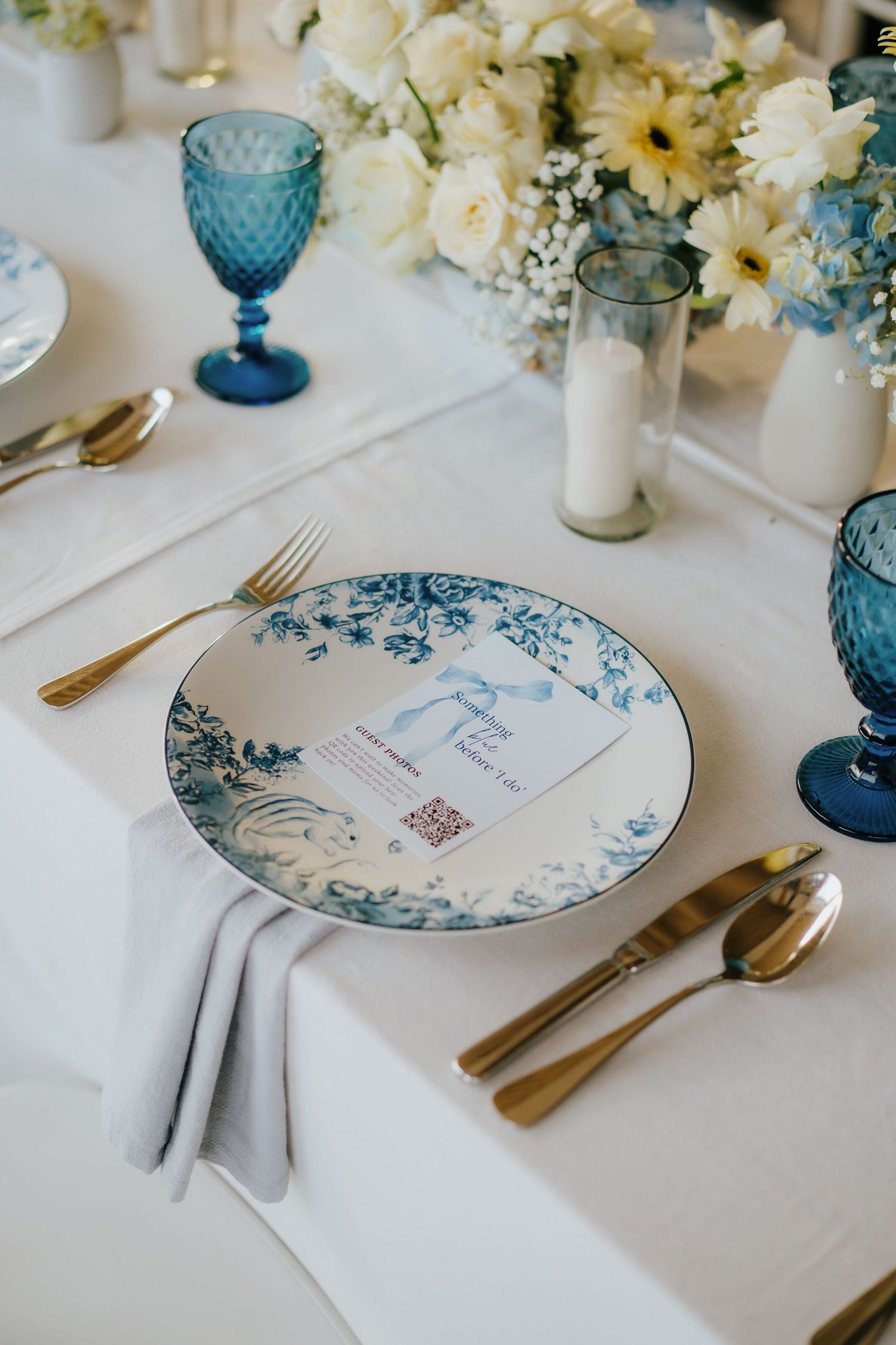 Elegant wedding table setting with blue toile plate, gold cutlery, blue goblets, and white floral centerpiece.