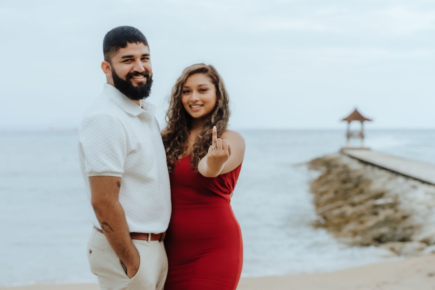 Couple in coordinating red and white outfits poses lovingly during a beach maternity photoshoot at sunset.