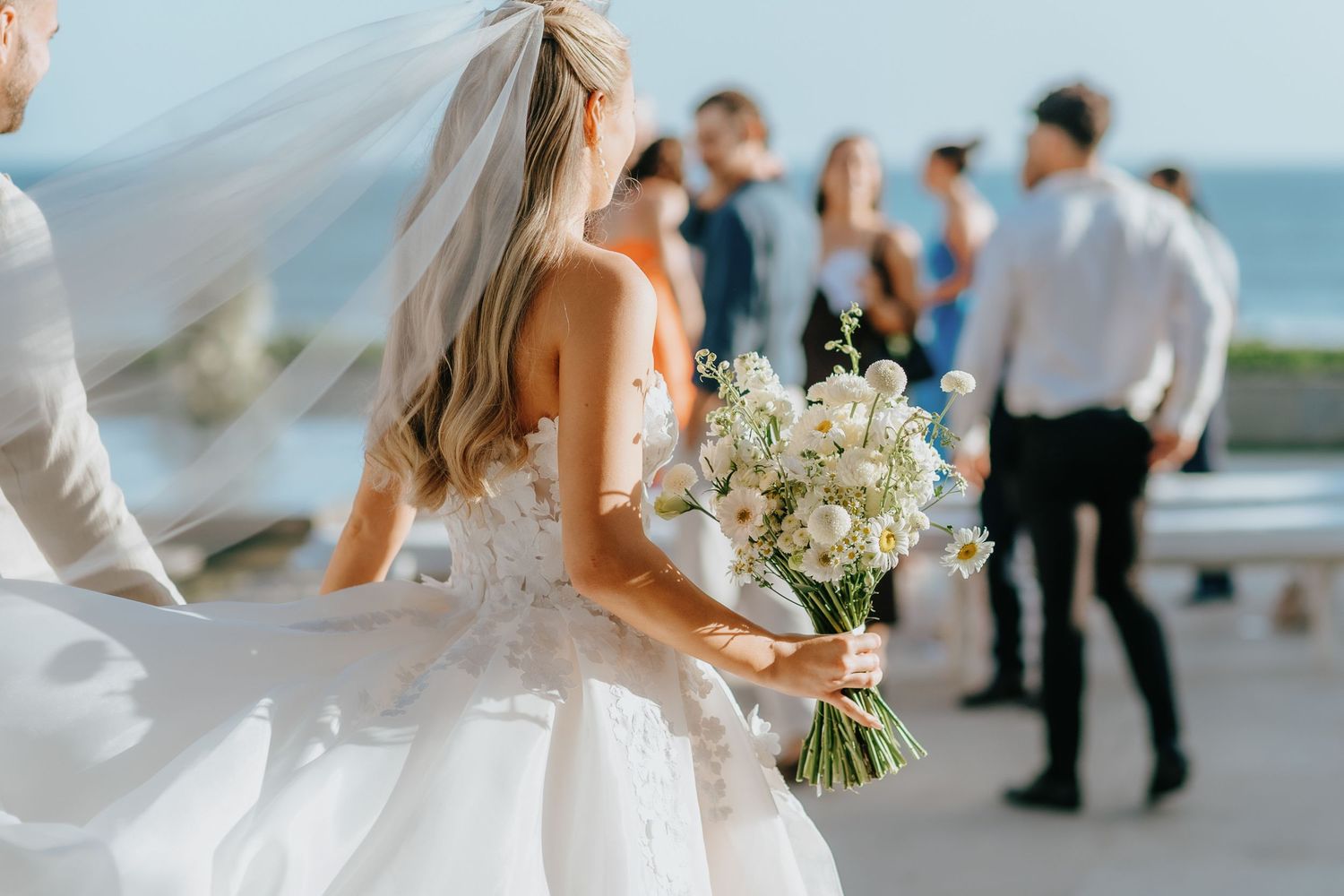Bride holding white floral bouquet in wedding dress with veil, guests mingling outdoors at sunny seaside ceremony.
