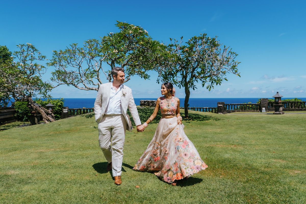 Couple in wedding attire walking hand-in-hand on lush green lawn with ocean view and tropical trees.