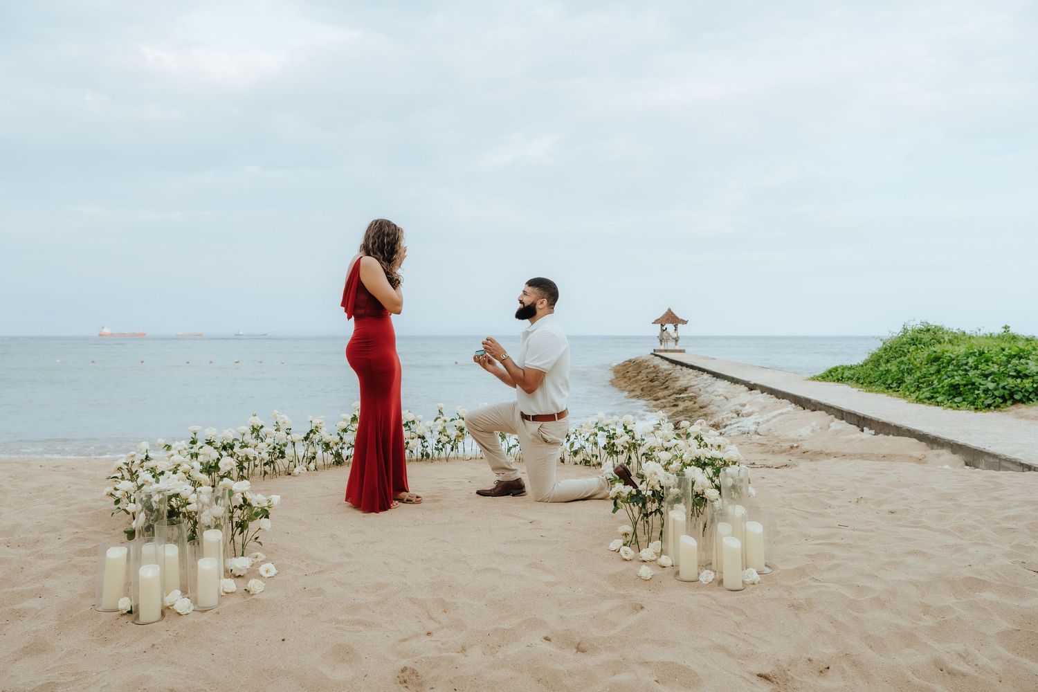 A romantic beach proposal setup with white floral arrangements and a couple sharing a special moment by the ocean.