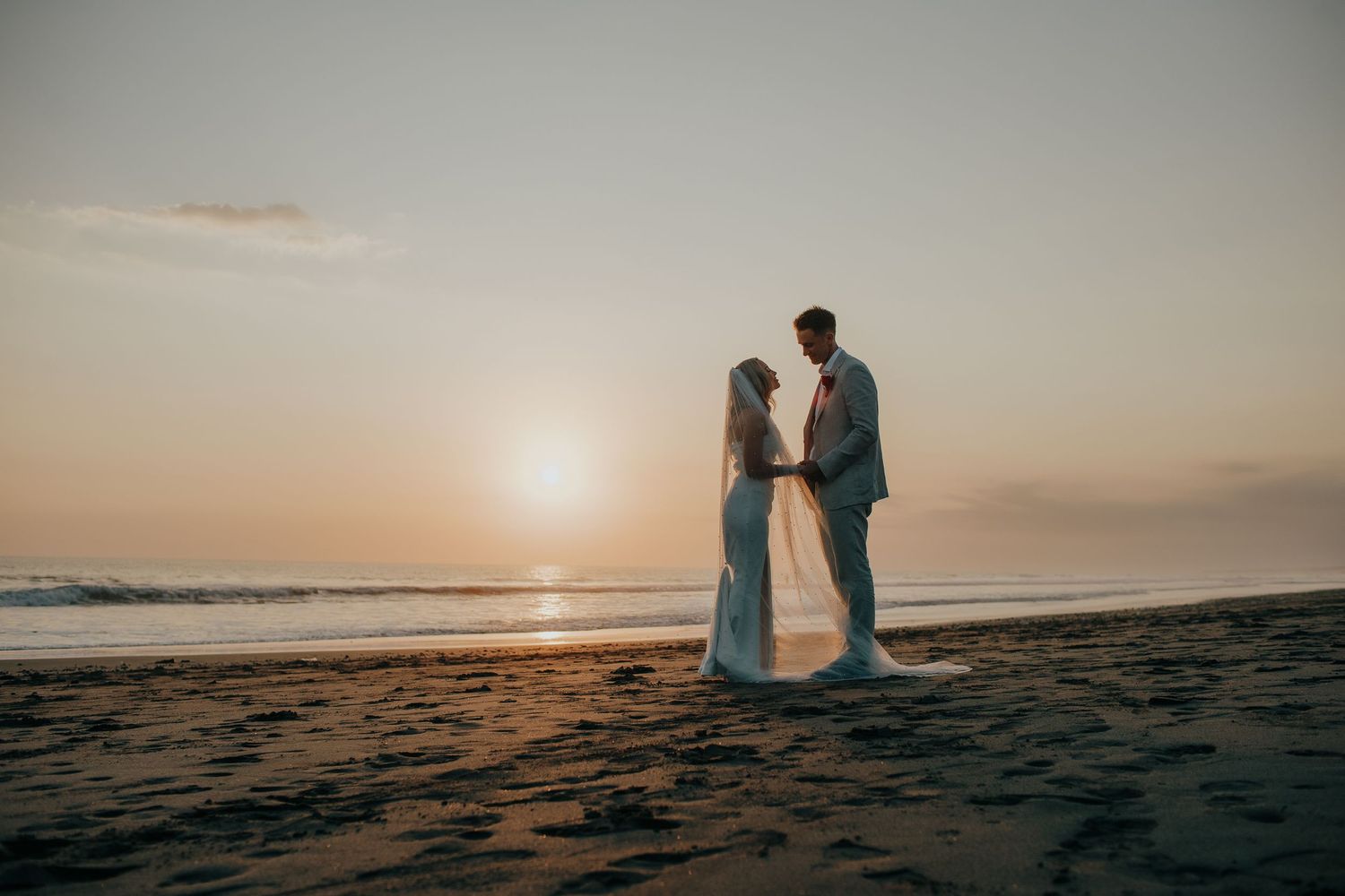 Couple holding hands on a sandy beach at sunset, bride in white gown with veil, groom in gray suit, ocean waves behind.