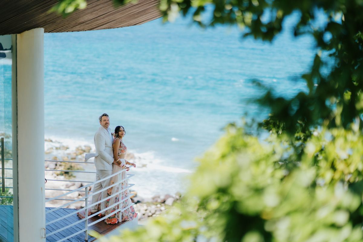 Couple stands on a seaside balcony with turquoise ocean waves and lush green trees framing a romantic coastal scene.