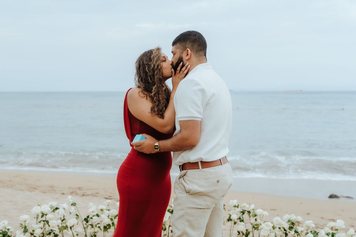 A romantic couple embraces on a sandy beach with ocean waves in the background.