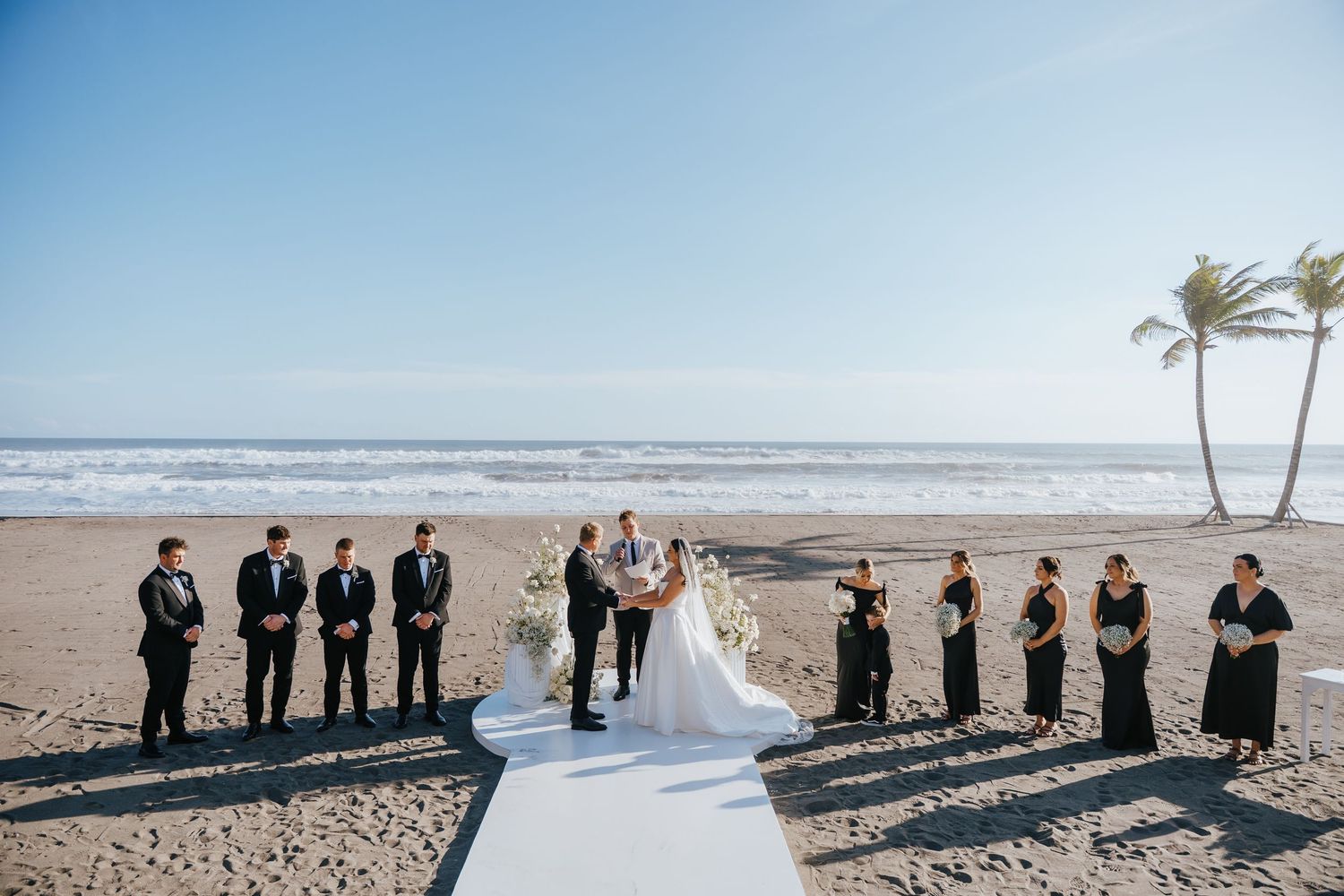 Elegant beach wedding ceremony with bride and groom at altar, groomsmen in black suits and bridesmaids in black dresses, ocean backdrop.