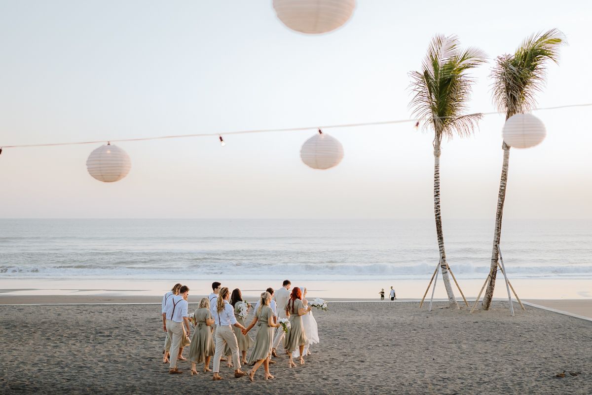 Wedding guests gather on a scenic beach at sunset with white paper lanterns decorating the ceremony space.