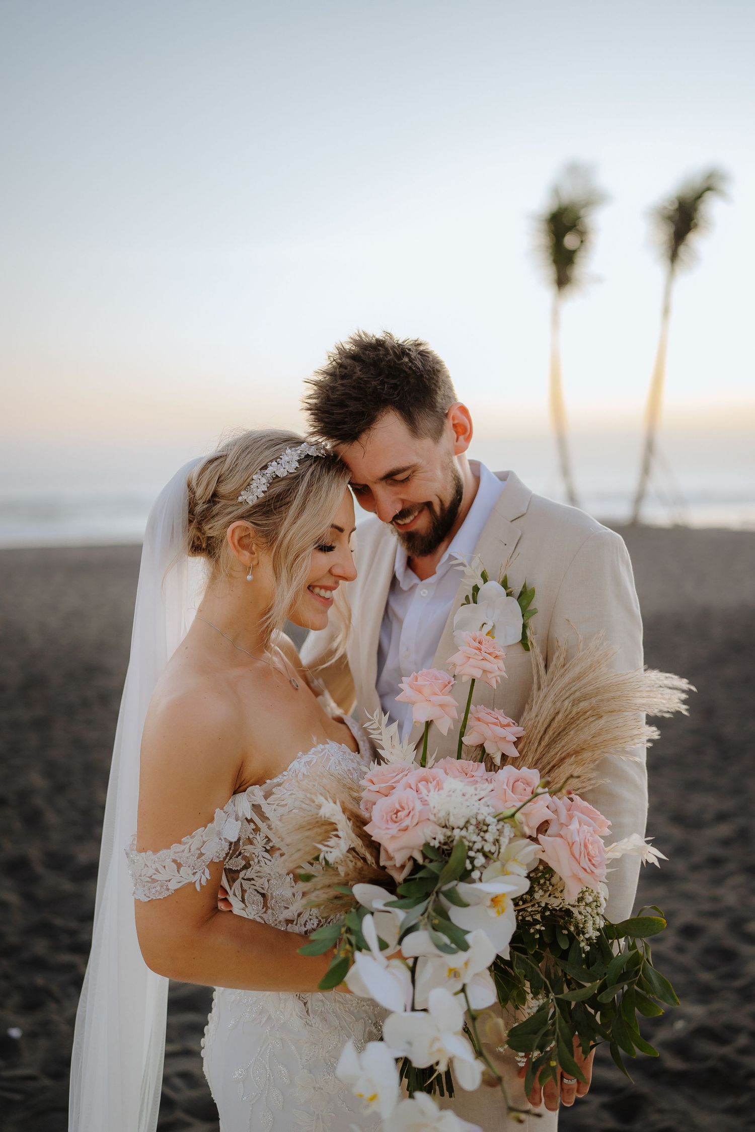 Bride and groom embrace on beach at sunset, she holds pink roses and orchid bouquet, wearing lace gown and veil.