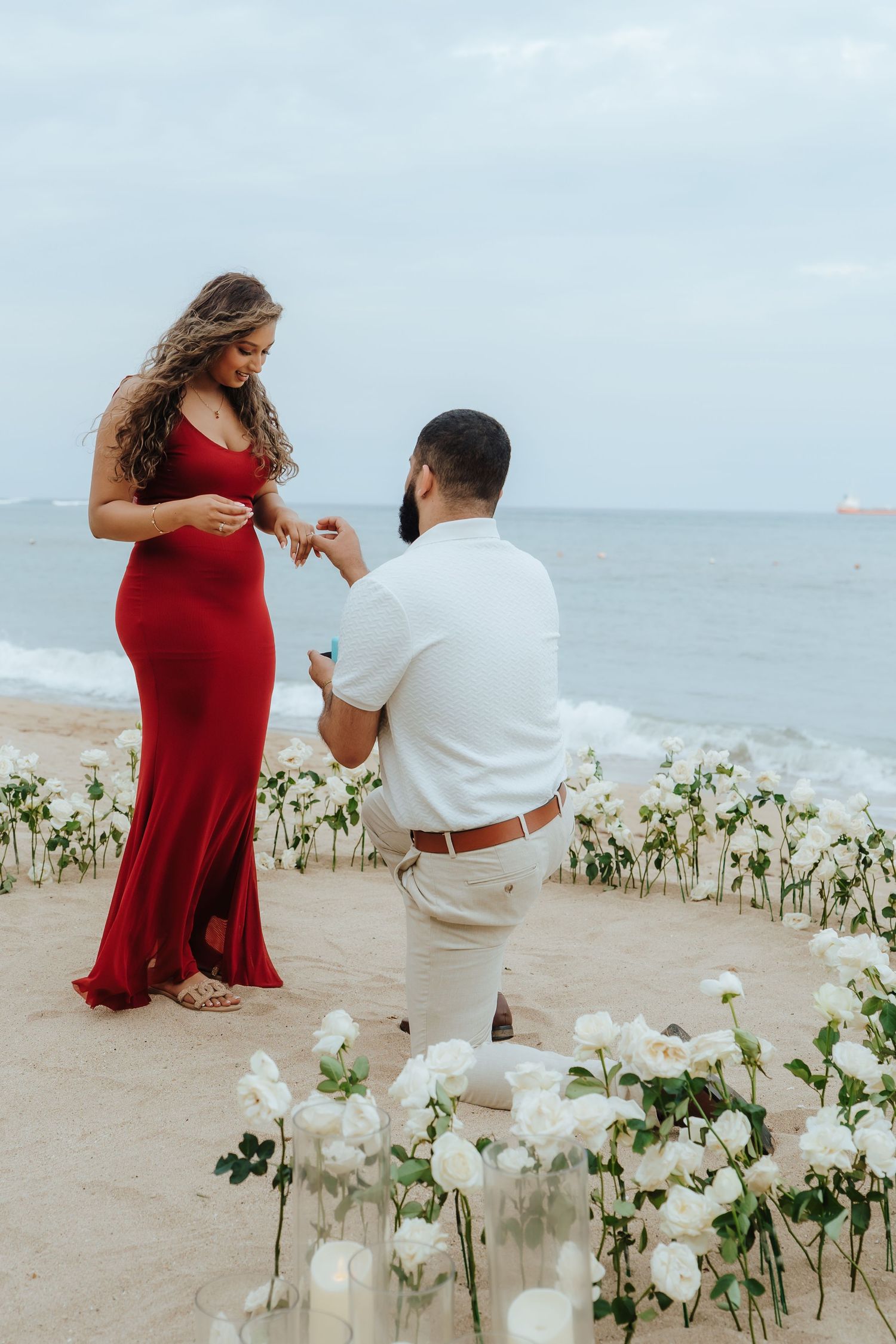 A couple shares a romantic proposal moment on the beach surrounded by white flowers and ocean views.