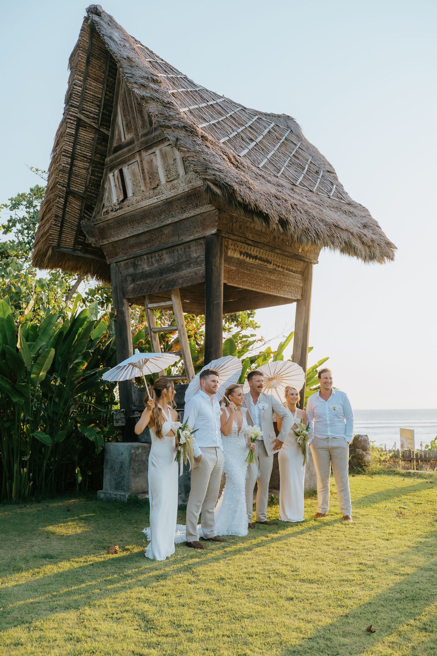 Wedding party in white attire holding parasols near a thatched bamboo hut with ocean views in Bali.