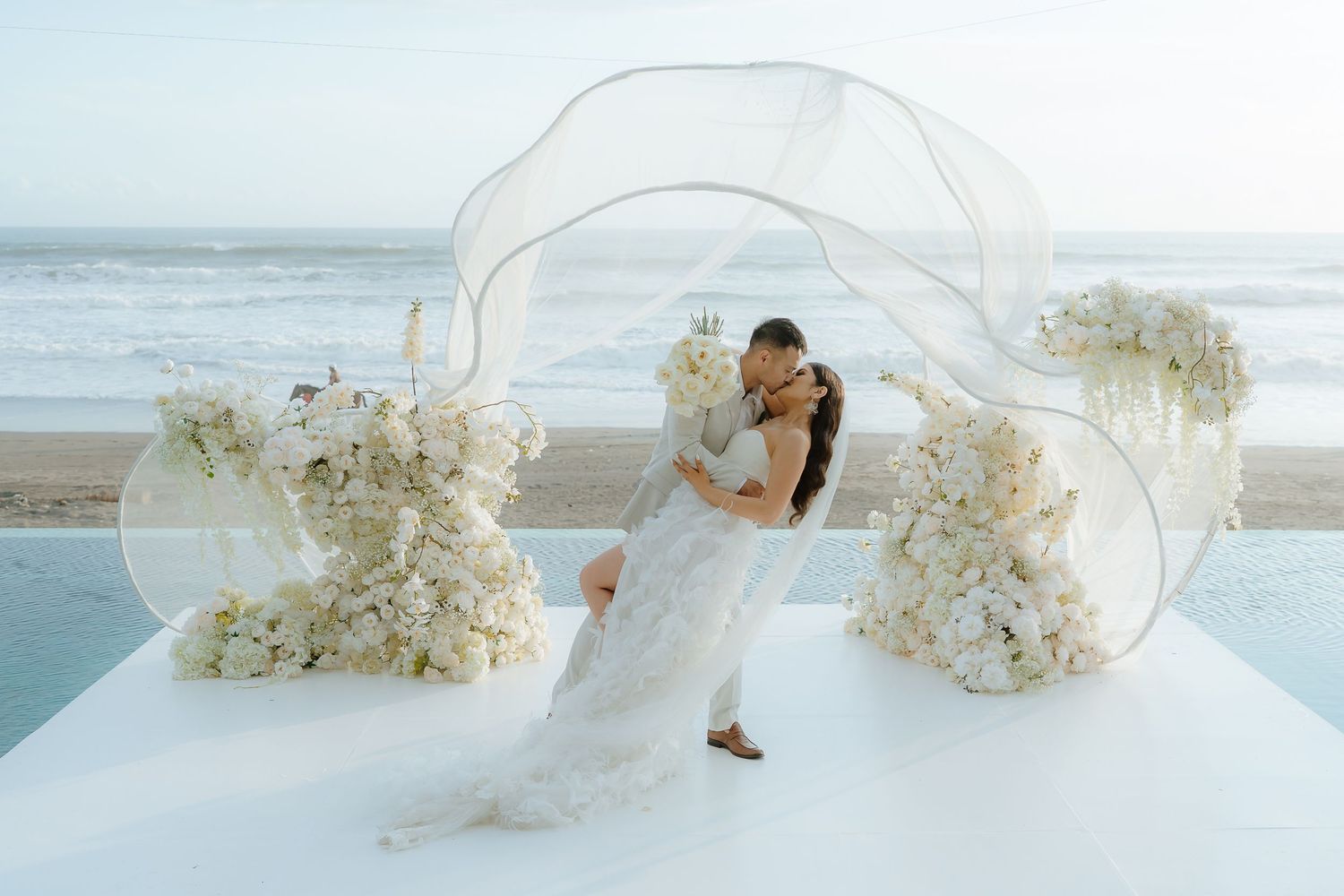 Bride and groom share romantic kiss under flowing veil at elegant beachside wedding ceremony with white floral arrangements.