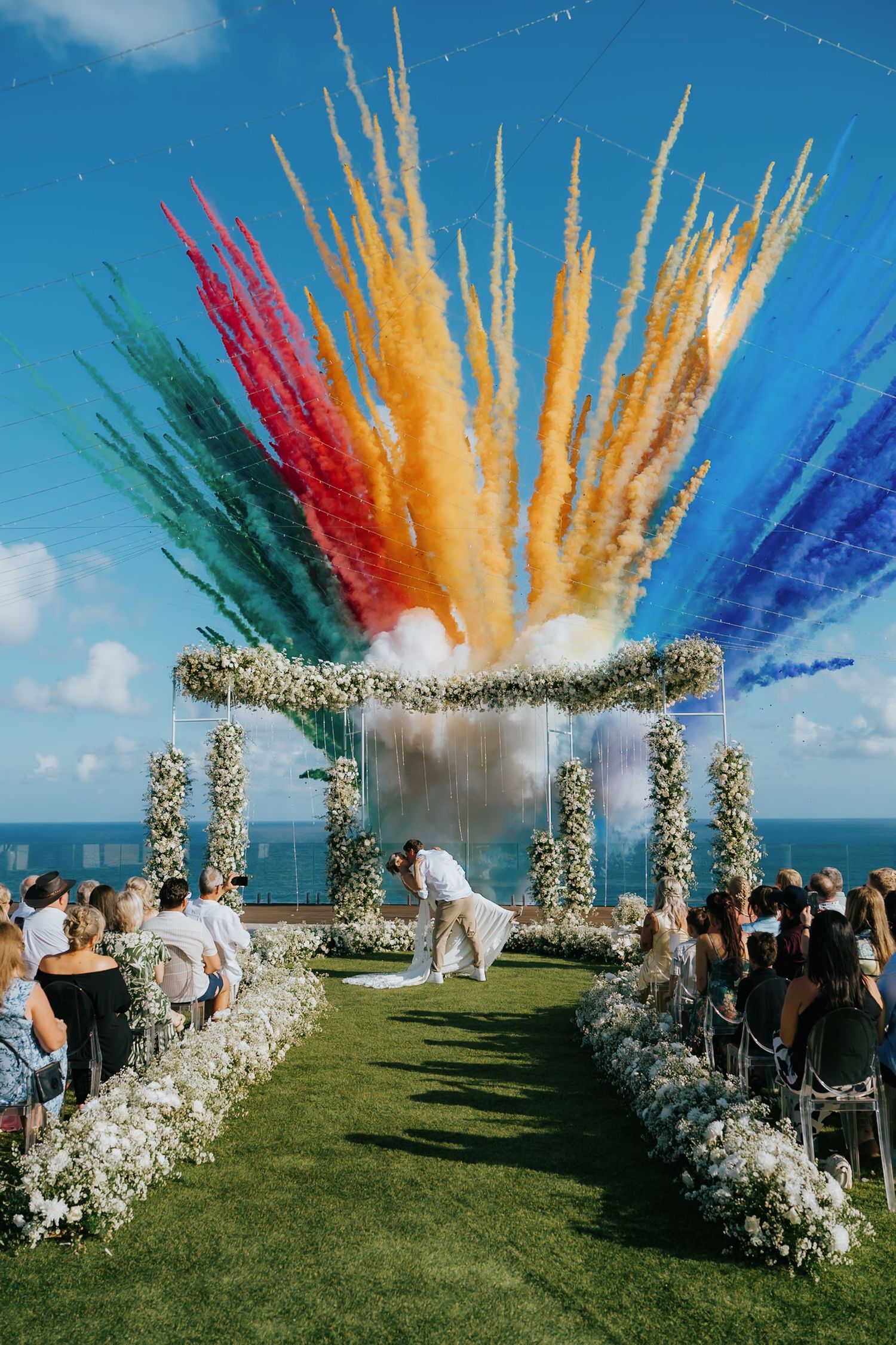 Colorful smoke bombs create vibrant rainbow explosion during outdoor wedding ceremony with floral arches by the ocean.