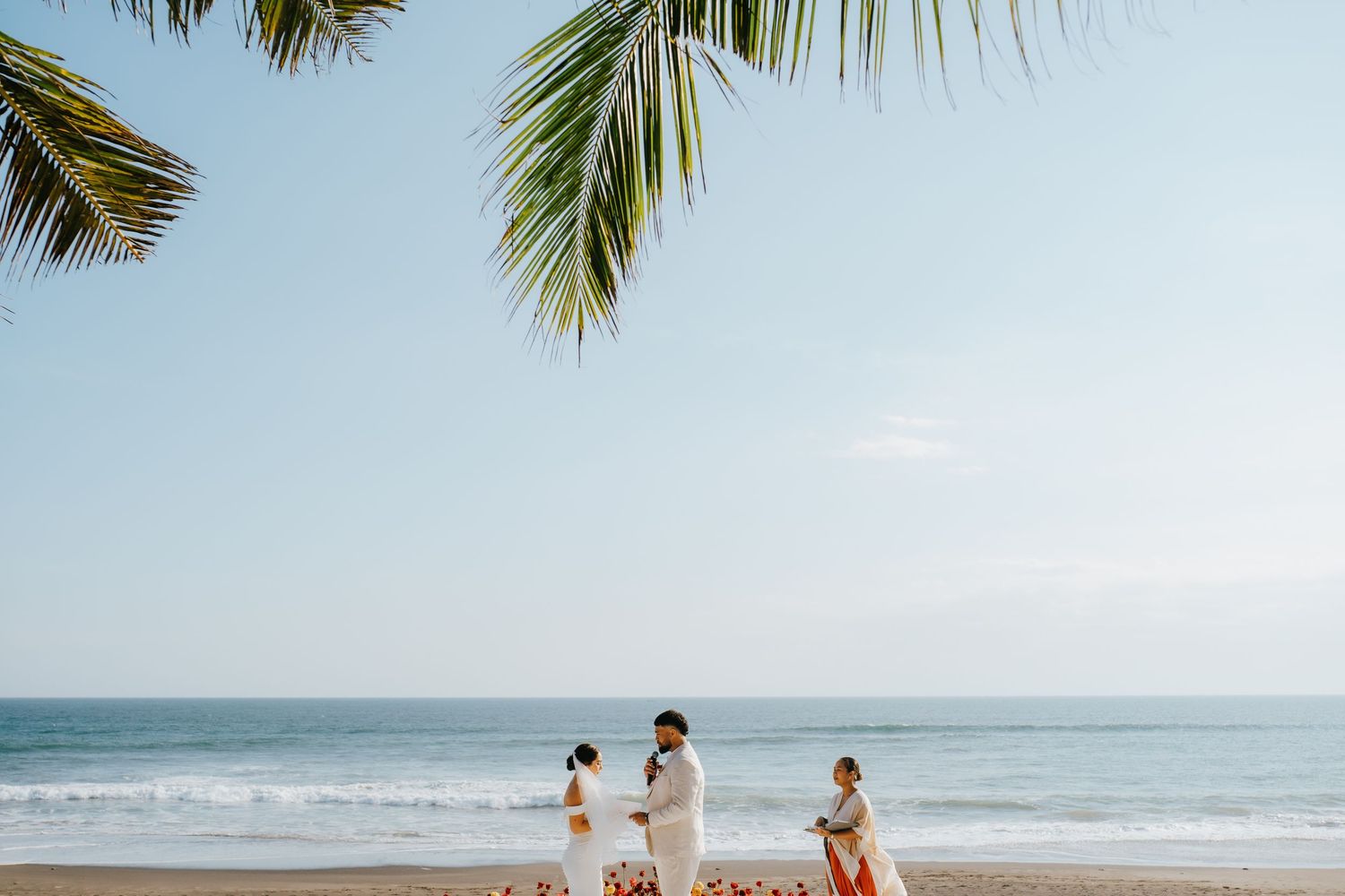 Couple exchanges vows on a tropical beach with palm trees and ocean backdrop during an intimate destination wedding ceremony.