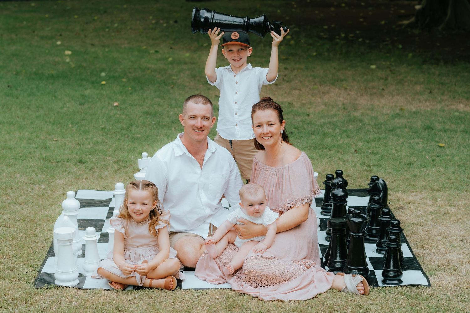 Family sitting on picnic blanket with chess pieces and camera equipment on grass.