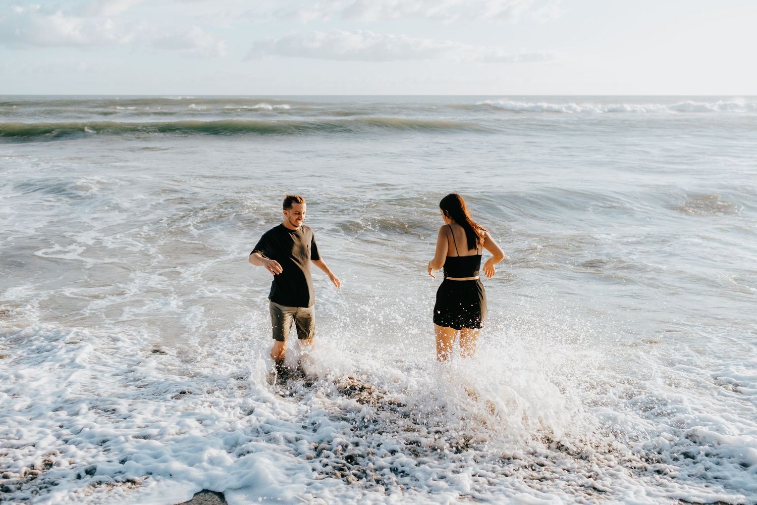 Two people wade through foamy ocean waves during a beach photoshoot.